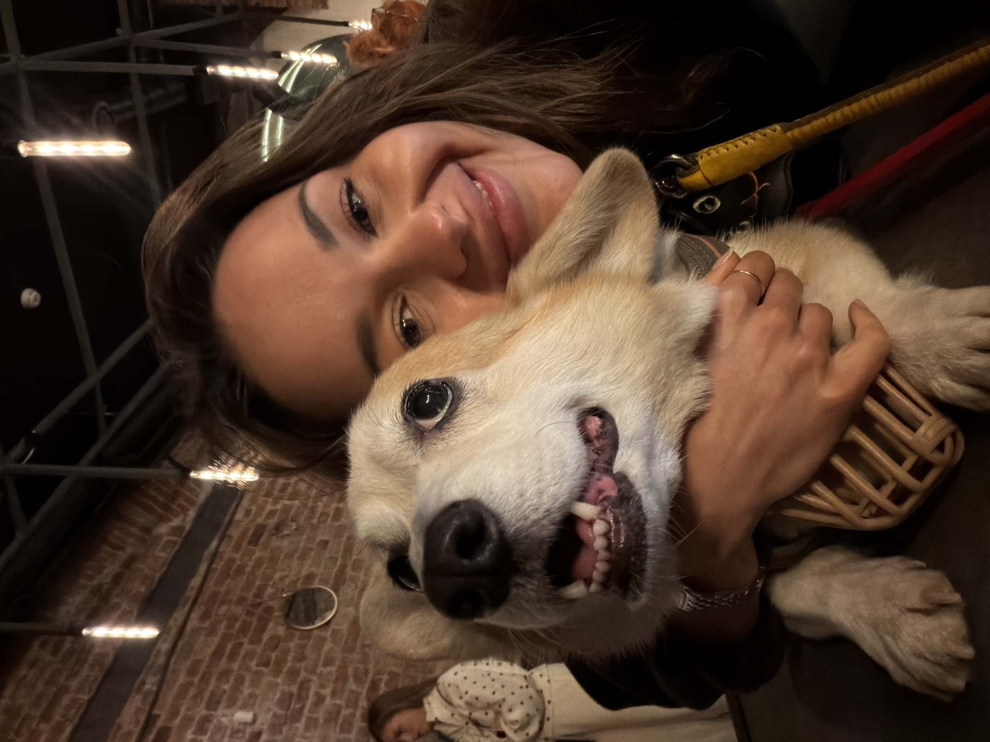 In Saint Petersburg, Russia, a woman and a dog are pictured in a close-up selfie. The woman has dark hair and is smiling warmly. She is embracing a light-colored corgi, whose head is close to the camera, with its mouth open, revealing its teeth and tongue. The dog appears to be wearing a muzzle. In the background, a brick wall and industrial-style lighting are visible, suggesting an indoor setting, possibly a restaurant or bar. Another person is partially visible in the background to the left. The overall atmosphere is casual and affectionate.