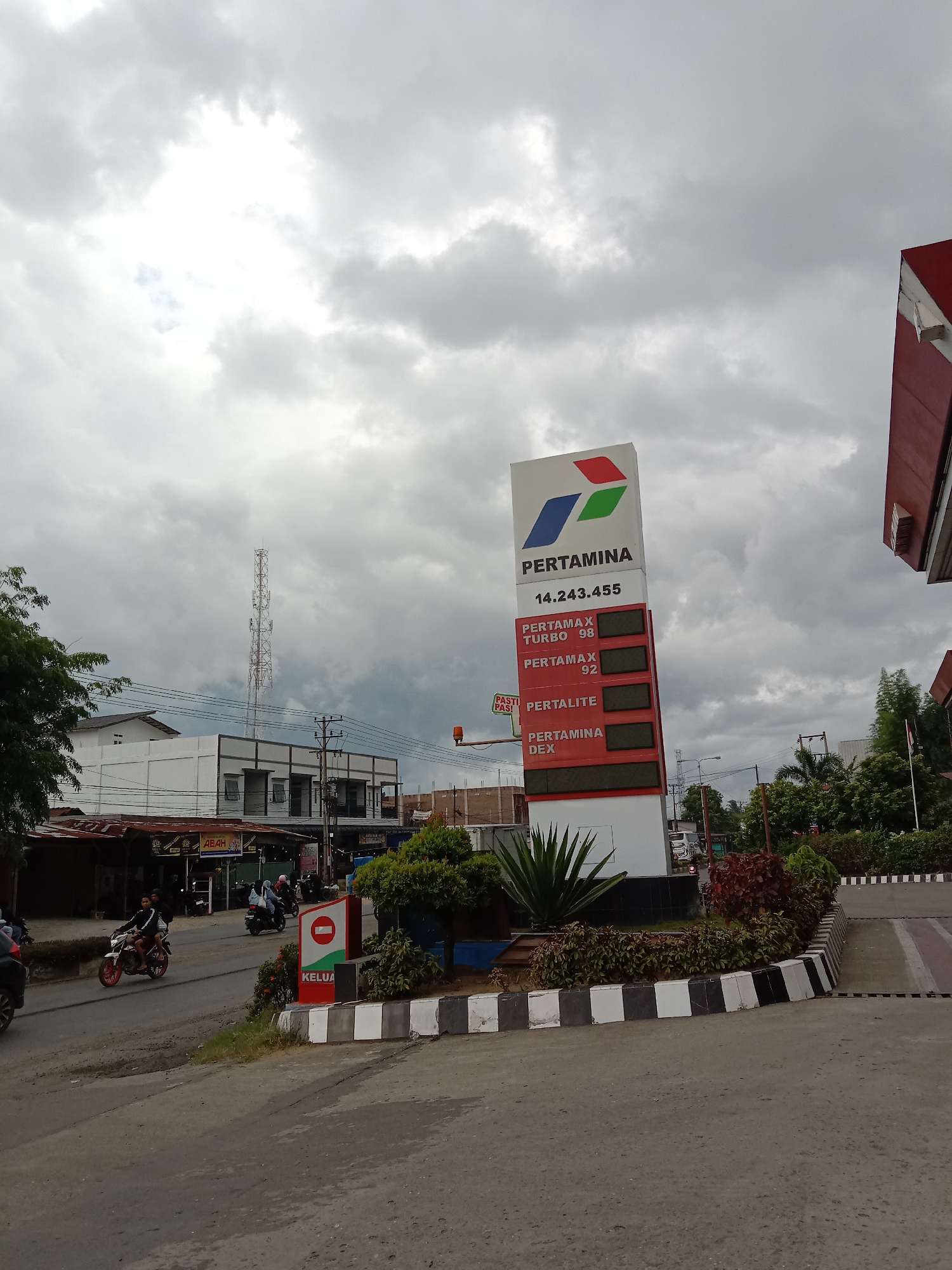 The image captures a scene at a Pertamina gas station in Lhokseumawe, Indonesia. The gas station is located on a street with other buildings and trees visible in the background. The sky is overcast with heavy clouds.
A prominent Pertamina sign displays the prices for various fuel types, including Pertamax Turbo 98, Pertamax 92, Pertalite, and Pertamina Dex. The listed price for Pertamax Turbo 98 is 14,243,455.
Several motorcycles are seen on the road near the gas station, suggesting traffic activity. The presence of a "Keluar" (Exit) sign indicates the location of an exit point from the gas station. A tall telecommunications tower is visible in the distance behind buildings and powerlines.