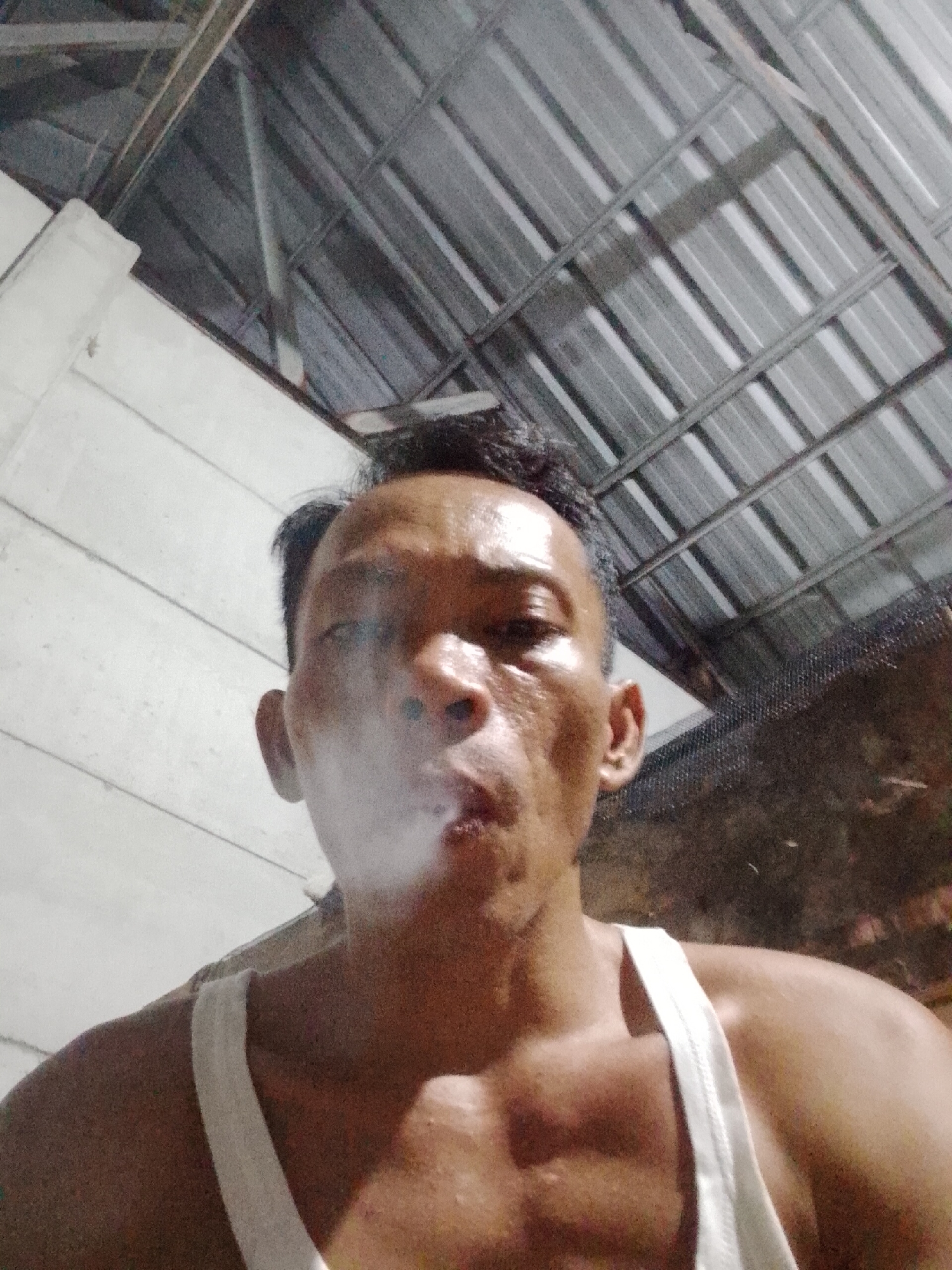A man wearing a white tank top is exhaling smoke, likely from a cigarette or vaping device. He appears to be indoors, possibly in a building with a corrugated metal roof. The location is suggested to be in Bukit Ketok, Indonesia. The lighting is dim. The man is the primary subject, and the smoke is a prominent element.