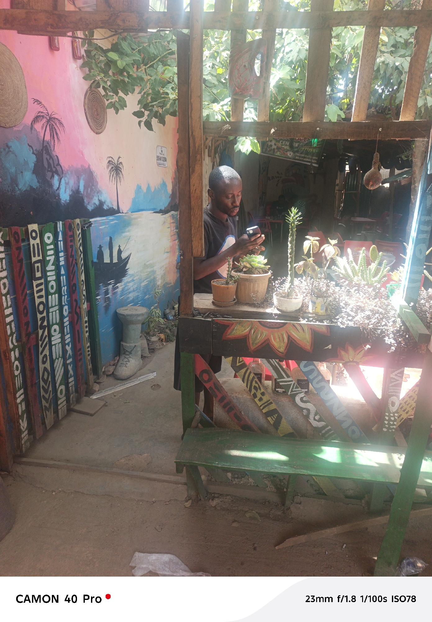 Here is a description of the image:
**Scene:** The image depicts an outdoor scene, likely a patio or courtyard, with a man standing behind a wooden structure. The scene includes various painted elements, plants, and seating, suggesting an artistic or bohemian setting.
**Location:** The presence of the phone number "5054110804" indicates a possible location within Nigeria, and the phone number format is typical for Nigeria. Without further context, a specific location within Jos cannot be definitively determined from the image.
**People:**
* A dark-skinned man is standing behind a decorated wooden counter, looking at a smartphone.
**Objects:**
* A wooden counter with potted plants, including cacti and succulents, is prominently featured.
* The counter is decorated with colorful paintings of flowers and abstract patterns.
* To the left, there are wooden boards with tribal-inspired patterns leaning against a wall.
* A mural depicting a seaside scene with palm trees and a boat is painted on the wall.
* A green bench is visible in the foreground.
* There is a concrete "shoe" planter near the painted boards.
**Actions:**
* The man is interacting with his smartphone.
**Environment:**
* The setting appears to be outdoors during the day. Bright sunlight is streaming through the wooden structure.
* The ground is dirt.
* The scene has a rustic, artistic feel, possibly indicating a cafe, art studio, or courtyard in a creative space.