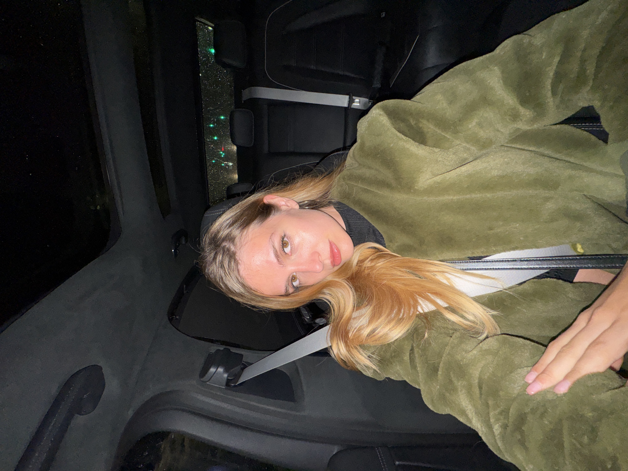 A young woman is reclining in the back seat of a car at night. The woman has light skin, shoulder-length blonde hair, and is wearing a green, plush jacket. She is also wearing a seatbelt. Outside the car window, blurred lights can be seen, suggestive of a city at night. The interior of the car is dark, with black leather seats and trim. The location is unknown.