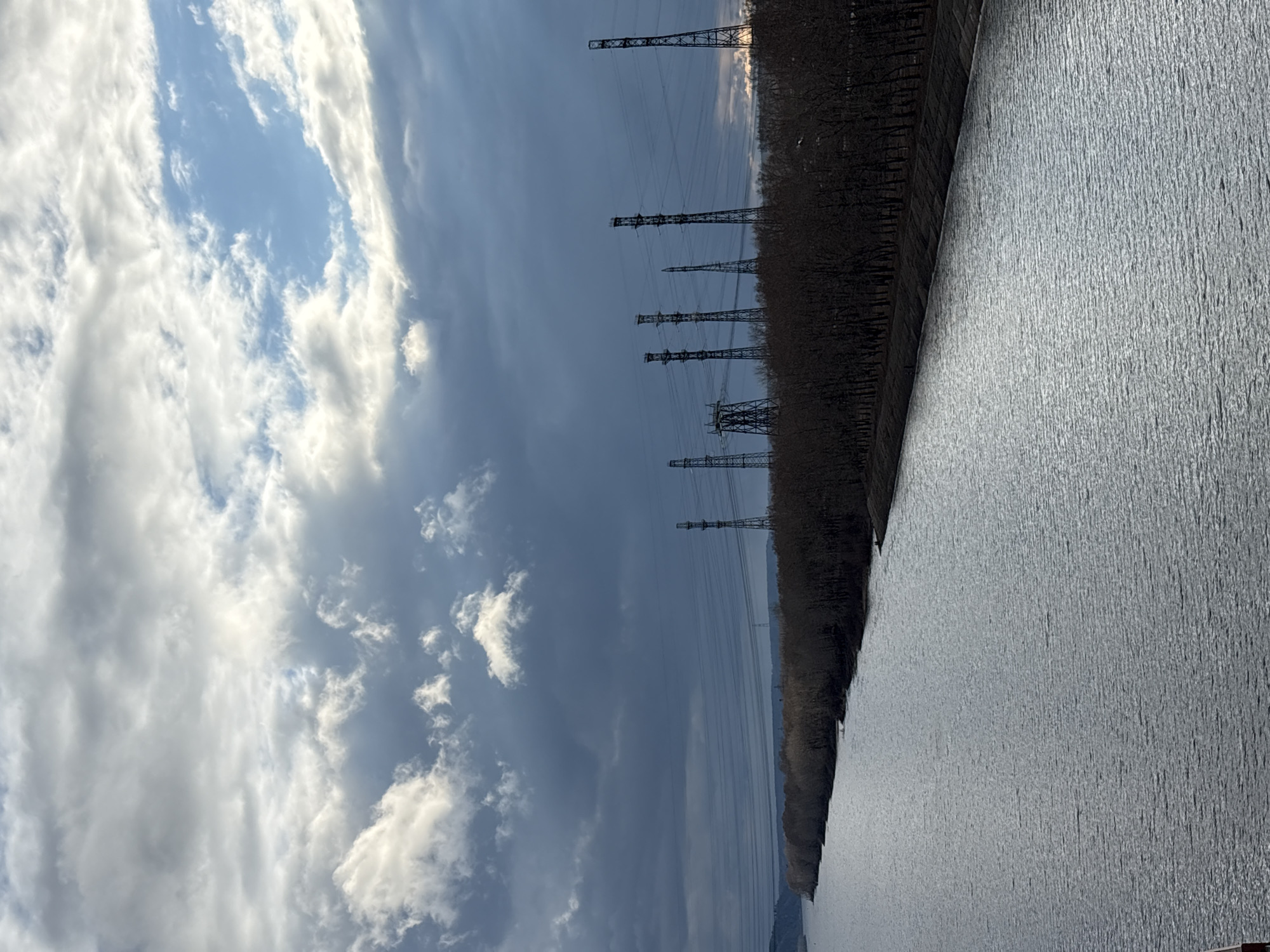 When viewed in its intended horizontal orientation, the image presents an expansive outdoor landscape. A wide body of water, likely a river or reservoir, dominates the lower and right portions of the frame, its surface appearing textured and shimmering under the daylight, reflecting light. Along the left bank, a dense, dark strip of land, possibly a forest of bare trees, runs into the distance. Numerous tall, lattice-structured power transmission towers are strategically placed along this bank, extending into the far background. The upper part of the image is filled with a dynamic sky, characterized by patches of clear blue interspersed with a variety of white and grey clouds, indicating partly cloudy weather. In the very far distance, a subtle outline of land or hills is visible beneath the horizon. The scene captures a tranquil daytime moment, blending natural elements with industrial infrastructure. There is no specific activity occurring, nor is any text visible. This setting is located in Chaikovsky, Russia, strongly suggesting the Kama River or Votkinsk Reservoir as the depicted body of water.