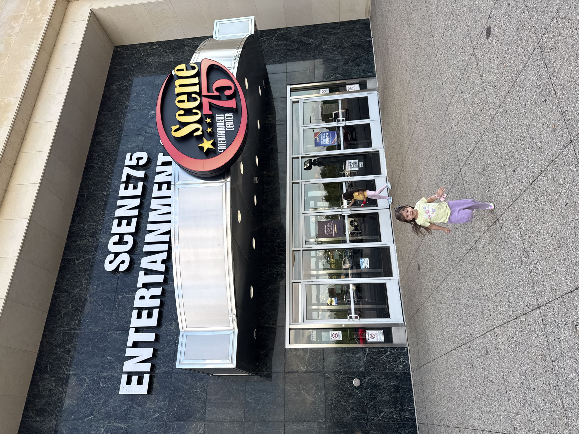Here is a detailed description of the image:
**Scene:**
The photo depicts the exterior of Scene75 Entertainment Center, a large entertainment venue. The building features a dark, possibly granite facade with bold white lettering spelling out "SCENE75 ENTERTAINMENT". A prominent, brightly colored sign is mounted above, displaying "Scene 75" with a red and black oval surrounding the text and three stars. The entrance is a series of glass doors framed in silver metal.
**People:**
A young girl is lying on the sidewalk in front of the entrance. She is wearing a yellow shirt and lavender pants. She is smiling and has one arm outstretched.
**Objects:**
* Scene75 Sign
* Glass Doors
* Sidewalk
* Building Facade
**Environment:**
The location is likely a commercial area due to the large entertainment center. The sidewalk appears to be made of small aggregate concrete. The building has a modern architectural style.
**Location:**
While not explicitly stated, Scene75 has a location in Columbus, Ohio, United States. Based on the architectural style and signage, it is highly probable that the image depicts the Columbus location.
