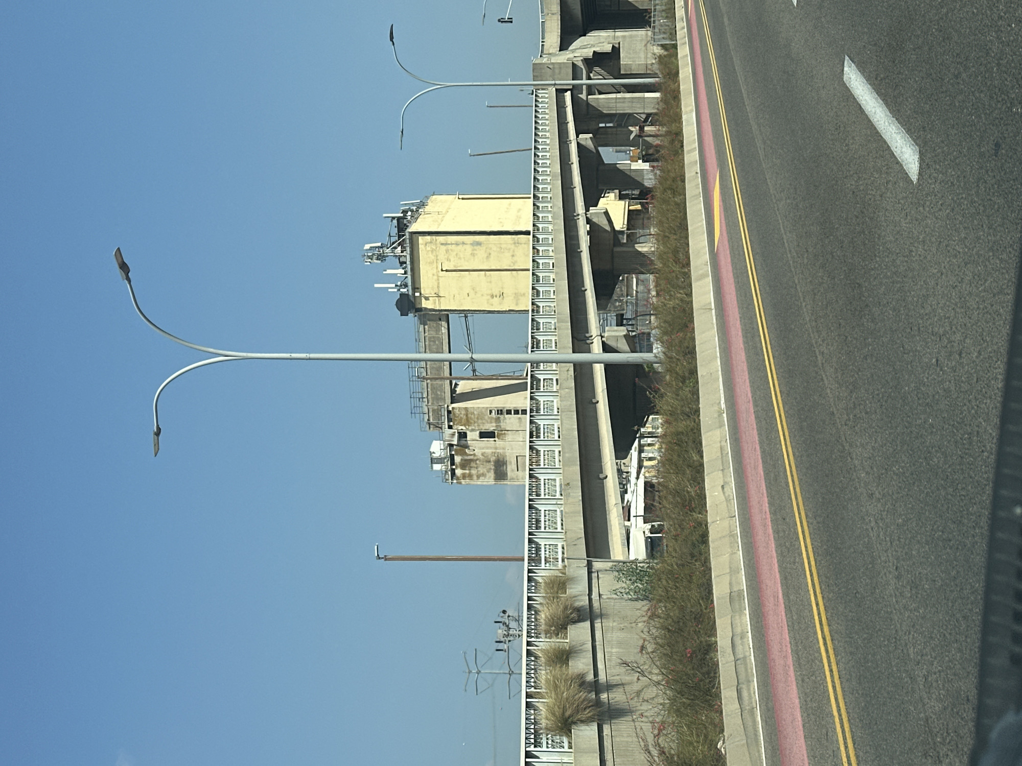 The image depicts a street-level view of a highway overpass in Haifa, Israel, on a clear, sunny day. The overpass, constructed from concrete, features a railing along its top edge, interspersed with decorative elements. Vegetation, including tall grasses and bushes, grows along the embankment beneath and alongside the road. Two prominent streetlights with curved arms are visible, one on the left and another on the right side of the image.

In the background, an old, yellow industrial building with a concrete structure and visible antennas on its roof stands behind the overpass. Further in the distance, a power line pylon and more industrial structures are discernible against the blue sky. The road in the foreground is asphalt, marked with yellow double lines and a pink lane. The overall scene suggests an urban or industrial setting. There are no people or vehicles immediately identifiable in the shot.