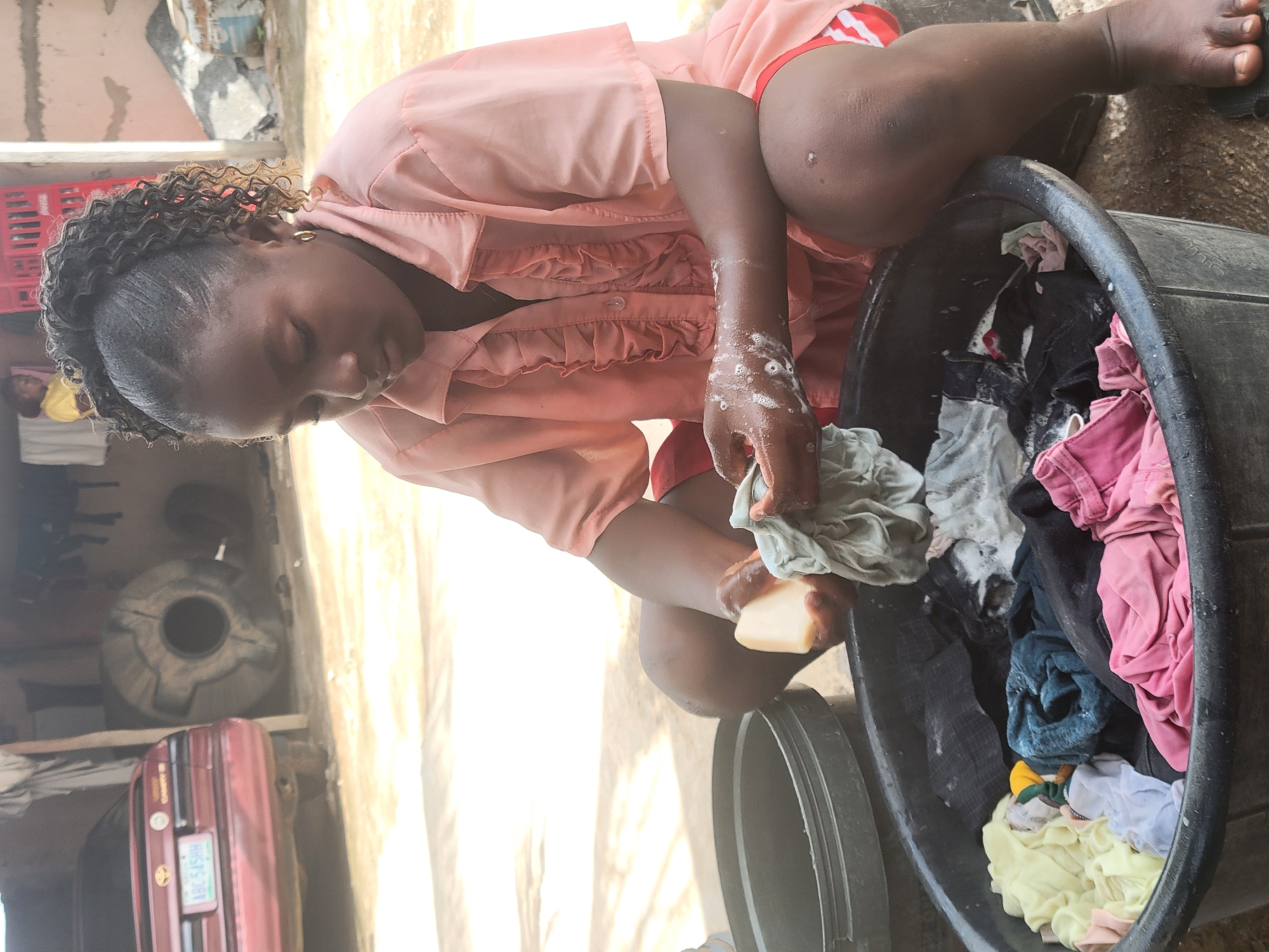 A woman washes clothes in a black plastic tub in Madalla, Nigeria. The woman, wearing a pink blouse and red shorts, kneels beside the tub, scrubbing a grey garment with a bar of soap. The tub contains various clothing items in pink, black, blue, and yellow. Behind the woman, there's a building, a red car, and a large cylindrical object.