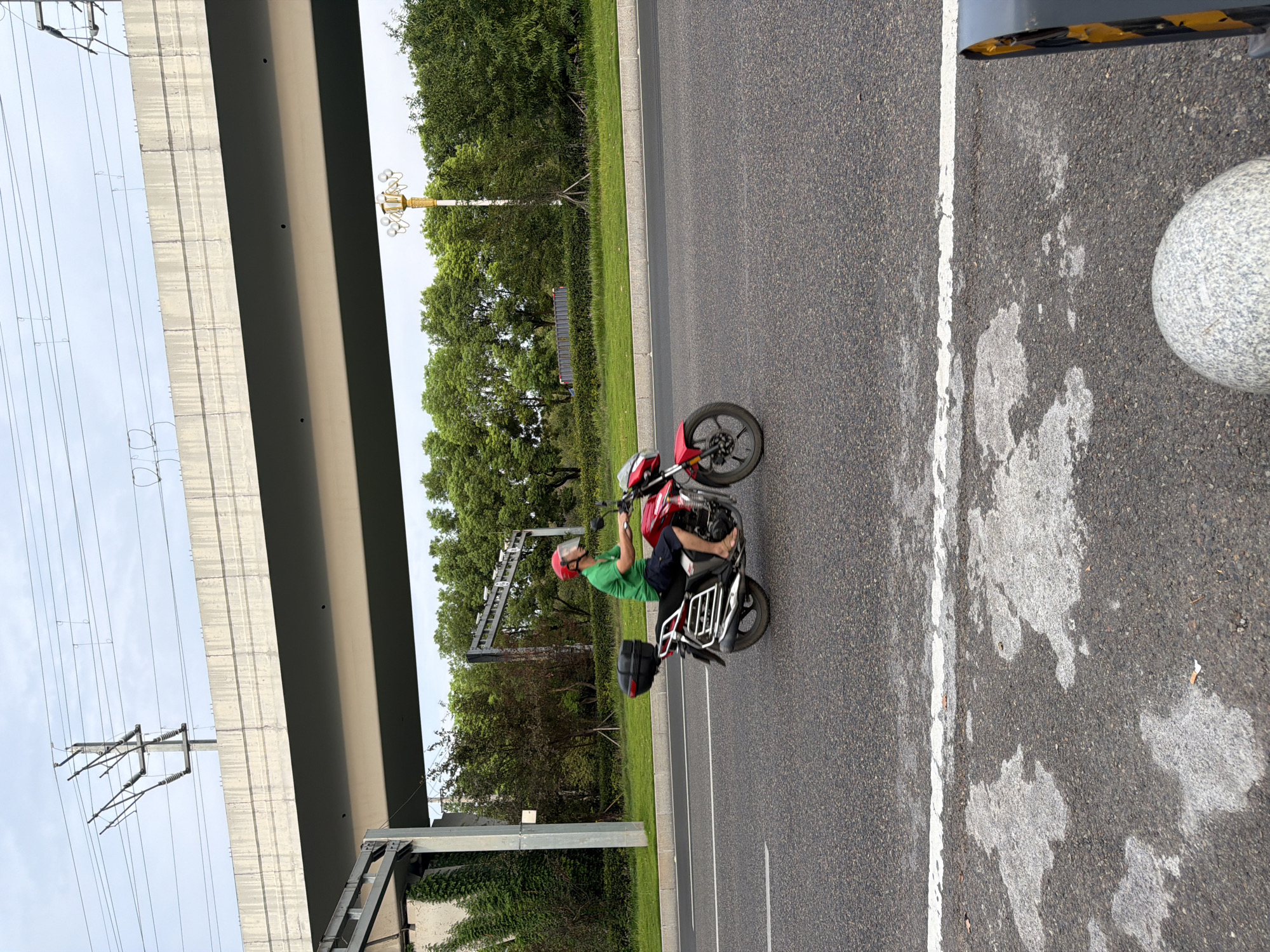 A person wearing a green shirt and red helmet rides a red motorcycle on a paved road in Jiangjin, China. The motorcycle has a black storage box on the back. Overhead, a concrete overpass with electrical lines is visible. Lush green trees line the roadside. The sky is overcast.