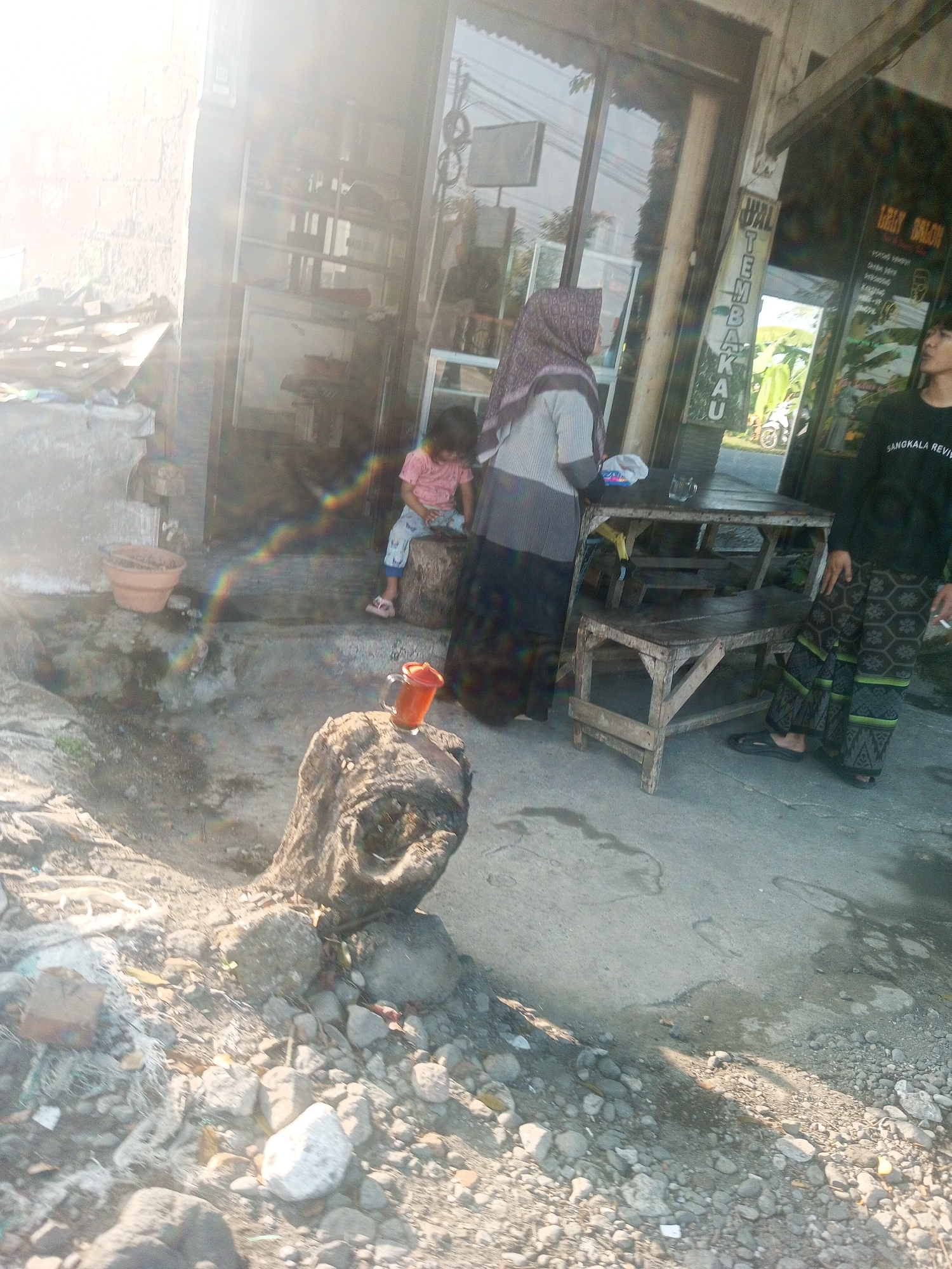 In the city of Krasak, Indonesia, a scene unfolds outside a shop with a glass storefront. The sun casts a bright glare, creating lens flare and obscuring some details.
In the foreground, a weathered, rough-textured tree stump stands, topped with a bright orange drinking mug. Scattered around the stump are various rocks and debris, including a piece of red brick and white stringy material.
To the left, a terracotta pot sits on the ground near a weathered wall.
Inside the shop's entrance, a young child wearing a pink t-shirt and light-colored pants sits on a wooden log.
To the right of the child, a woman wearing a headscarf and a grey and black striped long-sleeved shirt and skirt stands near a rustic wooden table and bench set. A glass is visible on the table.
Further to the right, another individual, likely a man, stands wearing a black long-sleeved shirt with white lettering and patterned trousers. He appears to be holding a cigarette. The shop sign behind him has text in a language that appears to be Indonesian, with the words "TEMBAKAU" visible vertically. Another sign above it appears to say "JUAL".
The ground surface is a mix of concrete and rough, stony terrain. The overall environment suggests an outdoor commercial area, possibly a small shop or market stall, with a utilitarian and somewhat unkempt appearance.