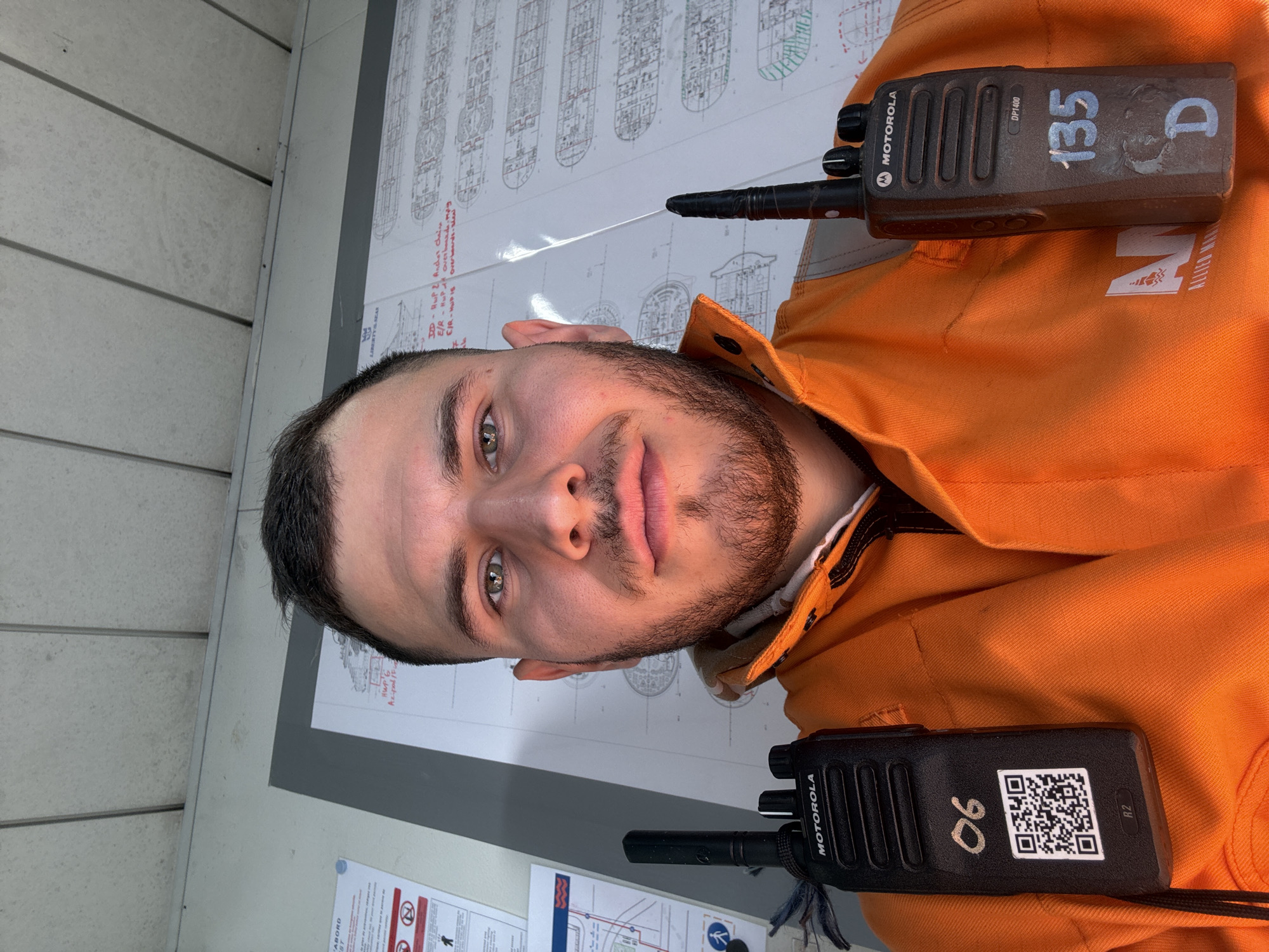 The image depicts a man wearing an orange work uniform with two Motorola DP1400 two-way radios, one labeled "135 D" and the other "06 R2". The man has short dark hair, a beard, and light-colored eyes. In the background, there is a map and blueprints of a ship or boat. There is also a sign with the word "ABORD" and another part reads "LIBERTY & SEAR". The phrase "Azipod/F" is visible as well. The environment suggests a maritime or industrial setting, possibly related to shipbuilding or port operations. The location can be inferred from clues on the map as Brest, France.