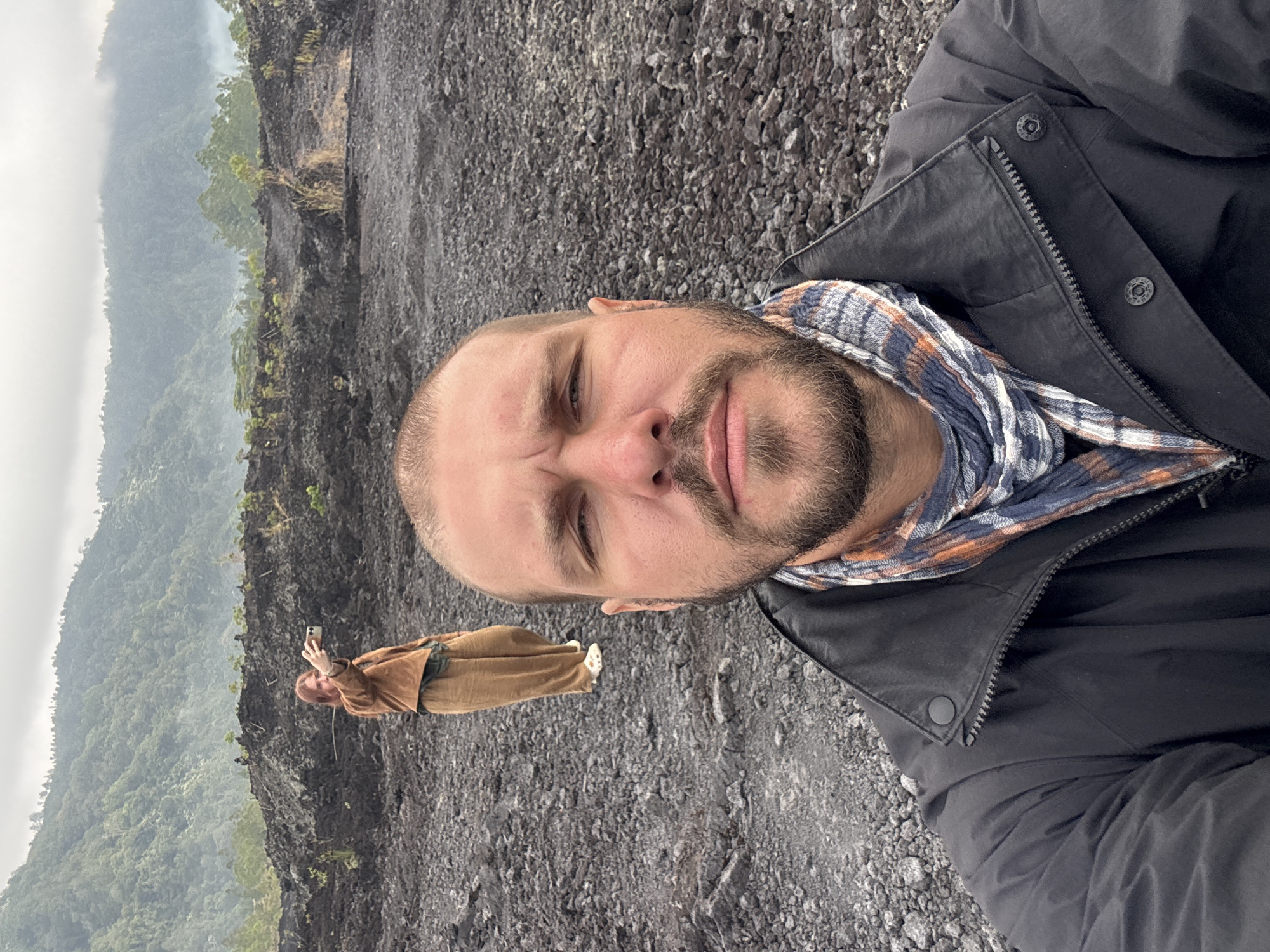 A man with a beard and short hair is taking a selfie in the foreground. He is wearing a black jacket and a checkered scarf. In the background, a woman with red hair is taking a photo with her phone. She is wearing a brown jacket and loose-fitting tan pants. The two individuals are standing on a rocky, dark-colored terrain that appears to be volcanic ash or lava rock. Behind them, lush green mountains rise up, partially shrouded in mist. The scene is set outdoors in a mountainous, potentially volcanic, landscape in Kintamani, Indonesia. The sky is overcast.