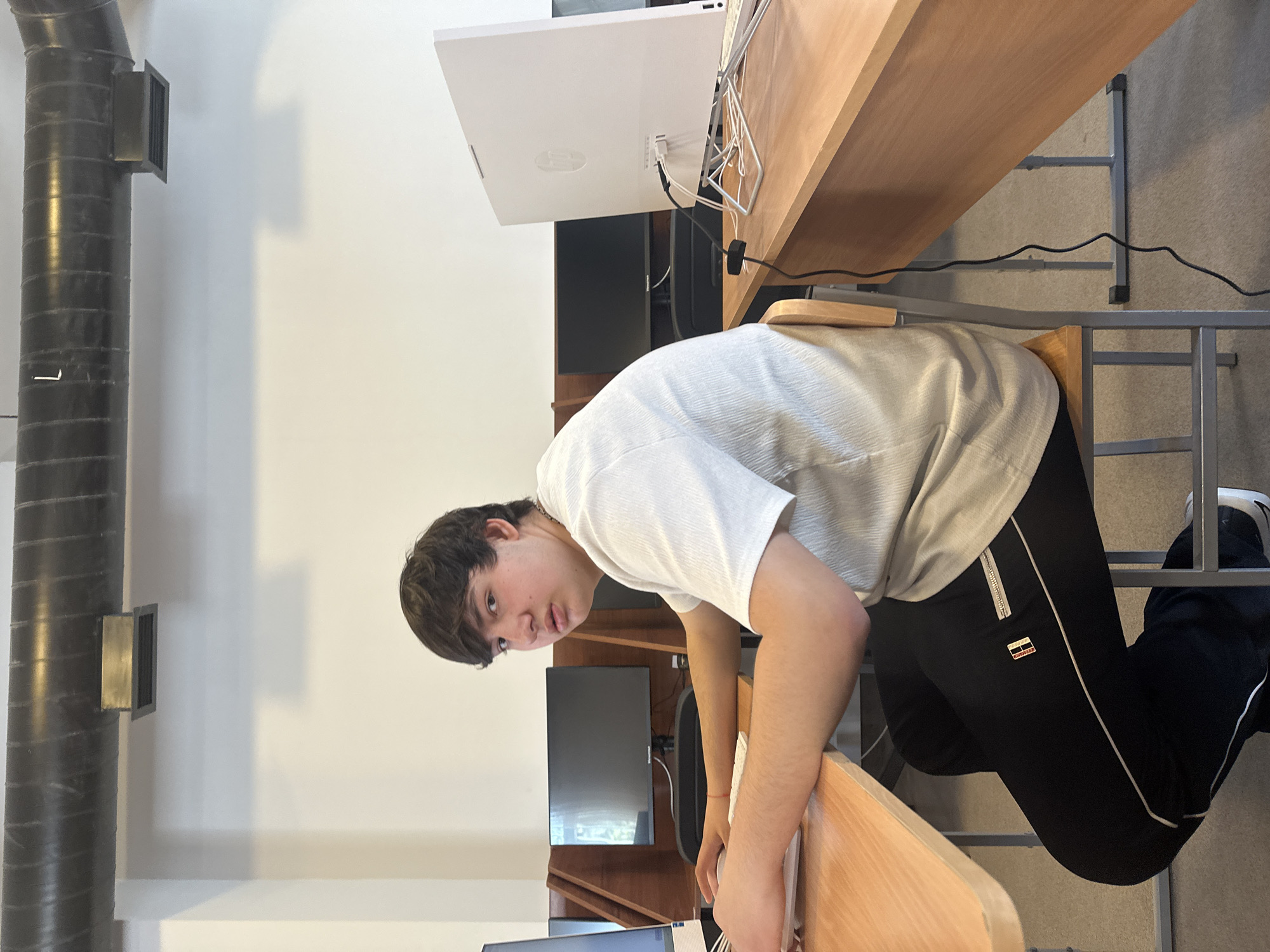 A young person with dark hair is seated at a desk in what appears to be a public computer lab. The person is wearing a light gray t-shirt and dark pants with white stripes. They are looking toward the camera with a neutral expression. On the desk in front of them is a white computer monitor. There are also other computers visible in the background.
The room has white walls with a dark, cylindrical structure running vertically along one wall. The walls and black pipe is indicative of the Tumo Center for Creative Technologies in Yerevan, Armenia.