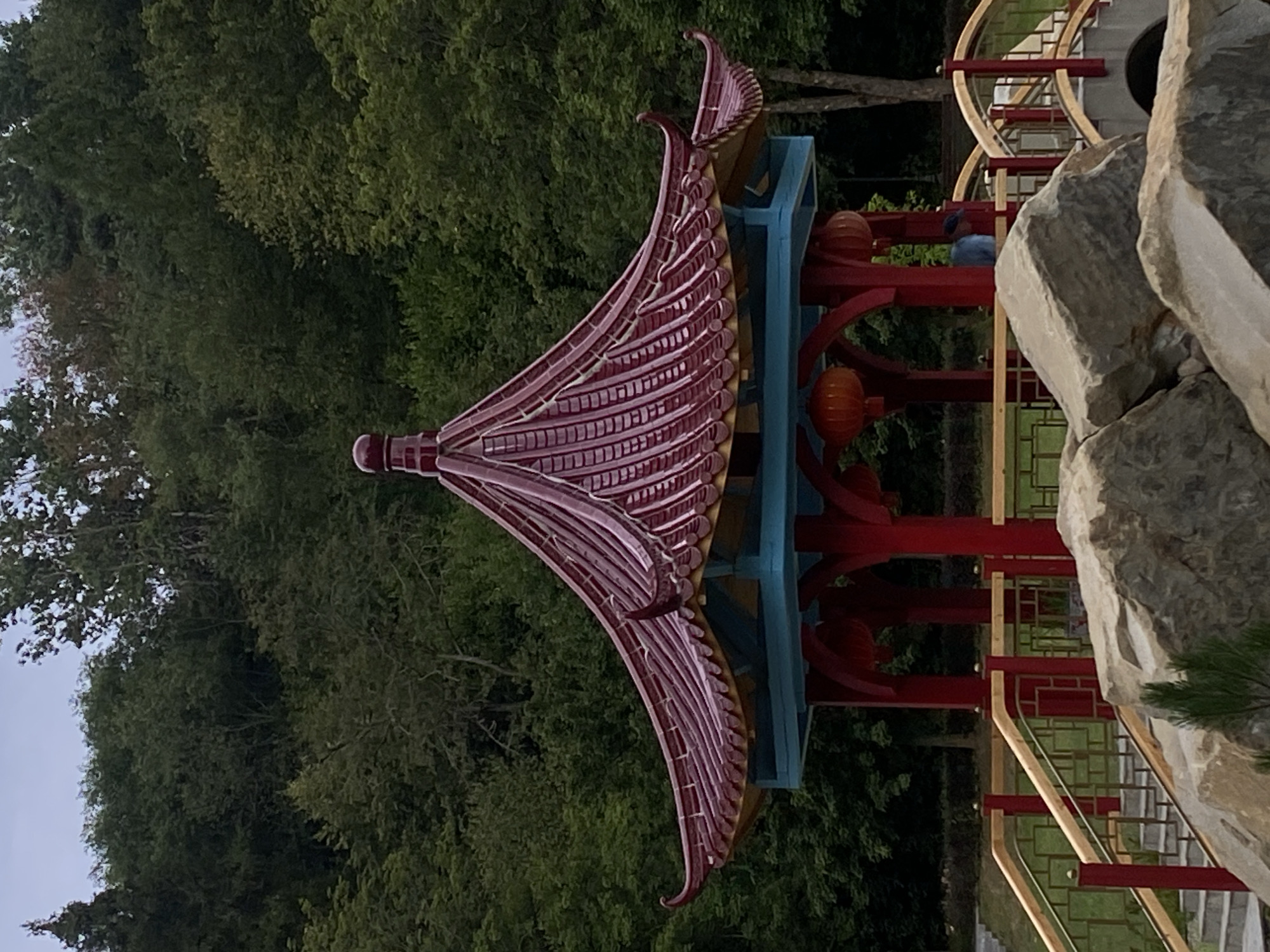 The image captures a traditional Chinese-style pavilion with a red, multi-tiered roof and blue accents, situated in a park-like setting. The pavilion is supported by red columns and features orange lanterns hanging from its structure. In front of the pavilion, there is a stone staircase with a red and gold railing, leading up to the structure. Large rocks are prominent in the foreground. The background consists of a dense forest with lush green trees. A person wearing a blue shirt and a dark cap is visible inside the pavilion. This scene is located in Muszyna, Poland, specifically in the Muszyna Japanese-style Garden.