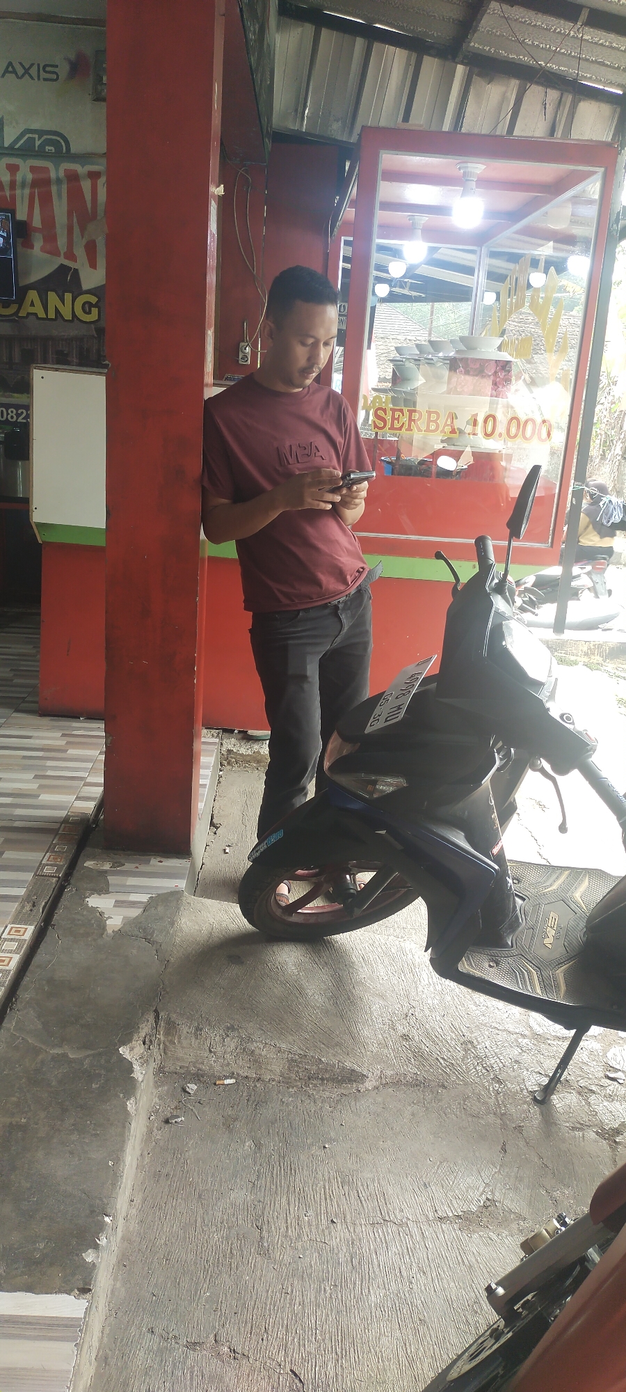 A young man stands leaning against a red pillar, looking at his phone. He is wearing a maroon t-shirt with a black design on the chest and black pants. In front of him is a dark-colored motorcycle. The man is positioned in what appears to be an outdoor area, possibly a shop entrance or a public space, with a building structure behind him. The ground is concrete, with some cracks and debris. A discarded cigarette butt is visible on the ground near the motorcycle's front tire. The background shows a glass display case with some items inside and signage, including text that reads "SERBA 10.000". The overall scene suggests a commercial or street-level setting in Cimaung, Indonesia.