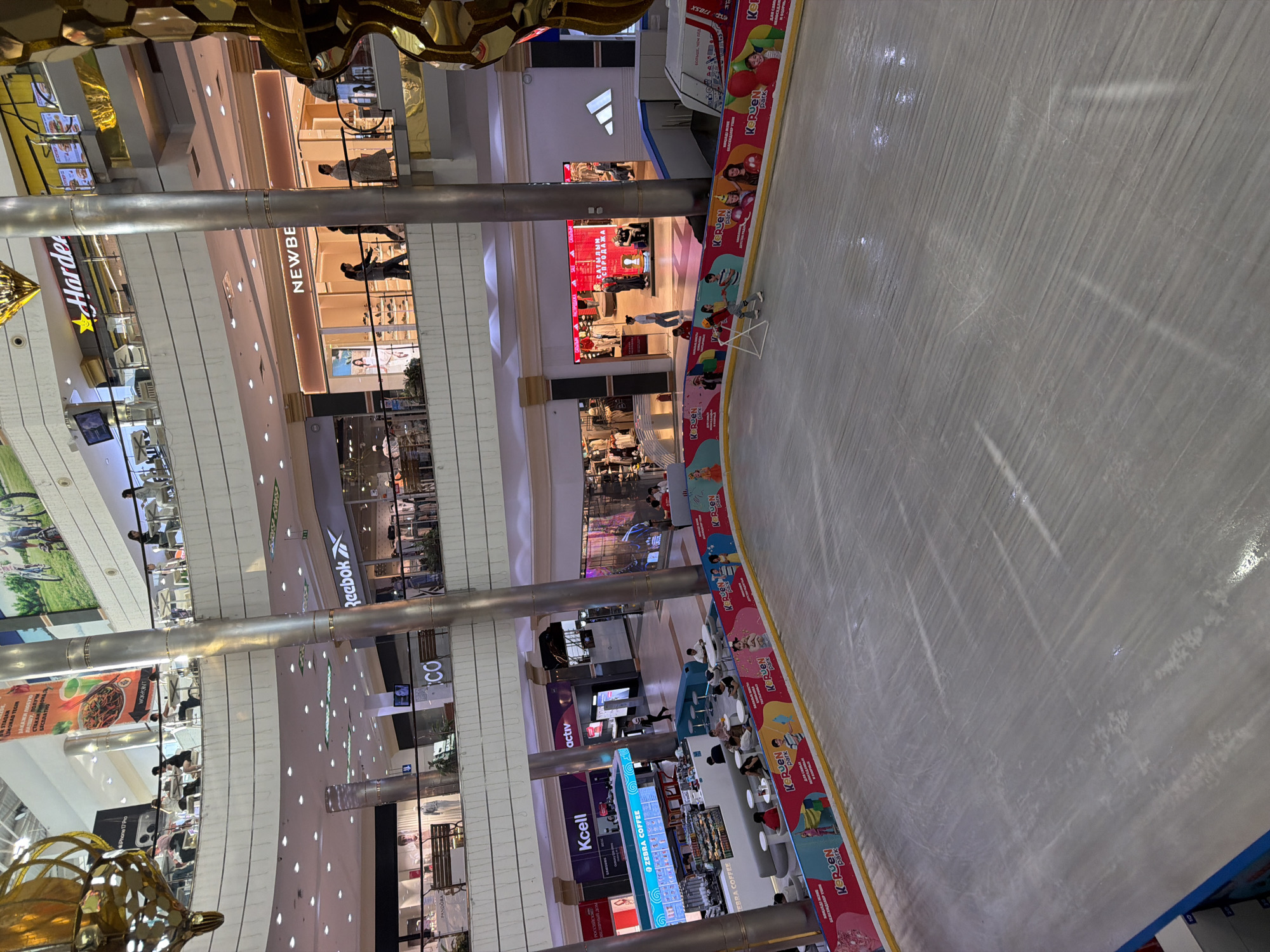 This is an interior view of a shopping mall in Aktobe, Kazakhstan, featuring an ice rink in the foreground. The ice rink is surrounded by a colorful barrier with advertisements for "KERUEN PARK". Several people are visible on and around the ice rink, including children and adults. One child is practicing skating with a training aid. In the background, multiple levels of the mall are visible, with shops, escalators, and food establishments. Notable shops include "Reebok", "NEWBEE", "Kcell", "Zebra Coffee", and "Hardee's". People are seen walking on the mall's walkways and sitting at tables. The mall appears to be well-lit with artificial lighting.