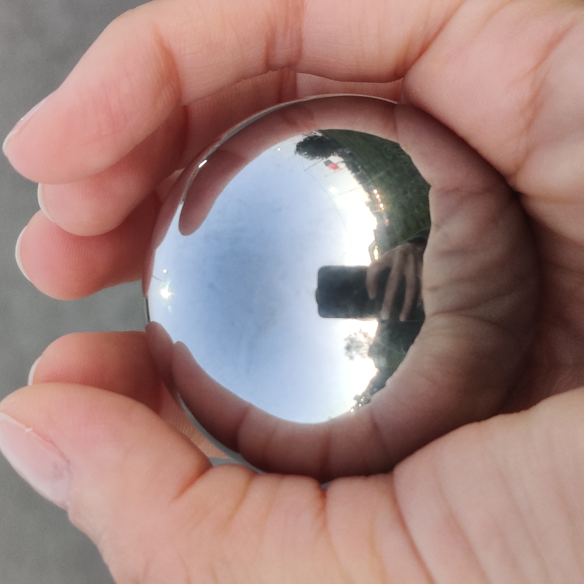 A close-up, eye-level shot shows a person's hands holding a polished metal sphere. The sphere acts as a convex mirror, reflecting a distorted, panoramic view of the surroundings. In the reflection, a black smartphone is visible, held by the same hands, indicating the image was taken by the person holding the sphere. The reflection also shows a sky with clouds, a grassy area, trees, and distant lights, possibly streetlights or car headlights, suggesting an outdoor urban setting during twilight or dawn. The overall impression is a personal, observational photograph using a reflective object. There is no specific information in the image that would allow for the identification of the city as Yekaterinburg, Russia.