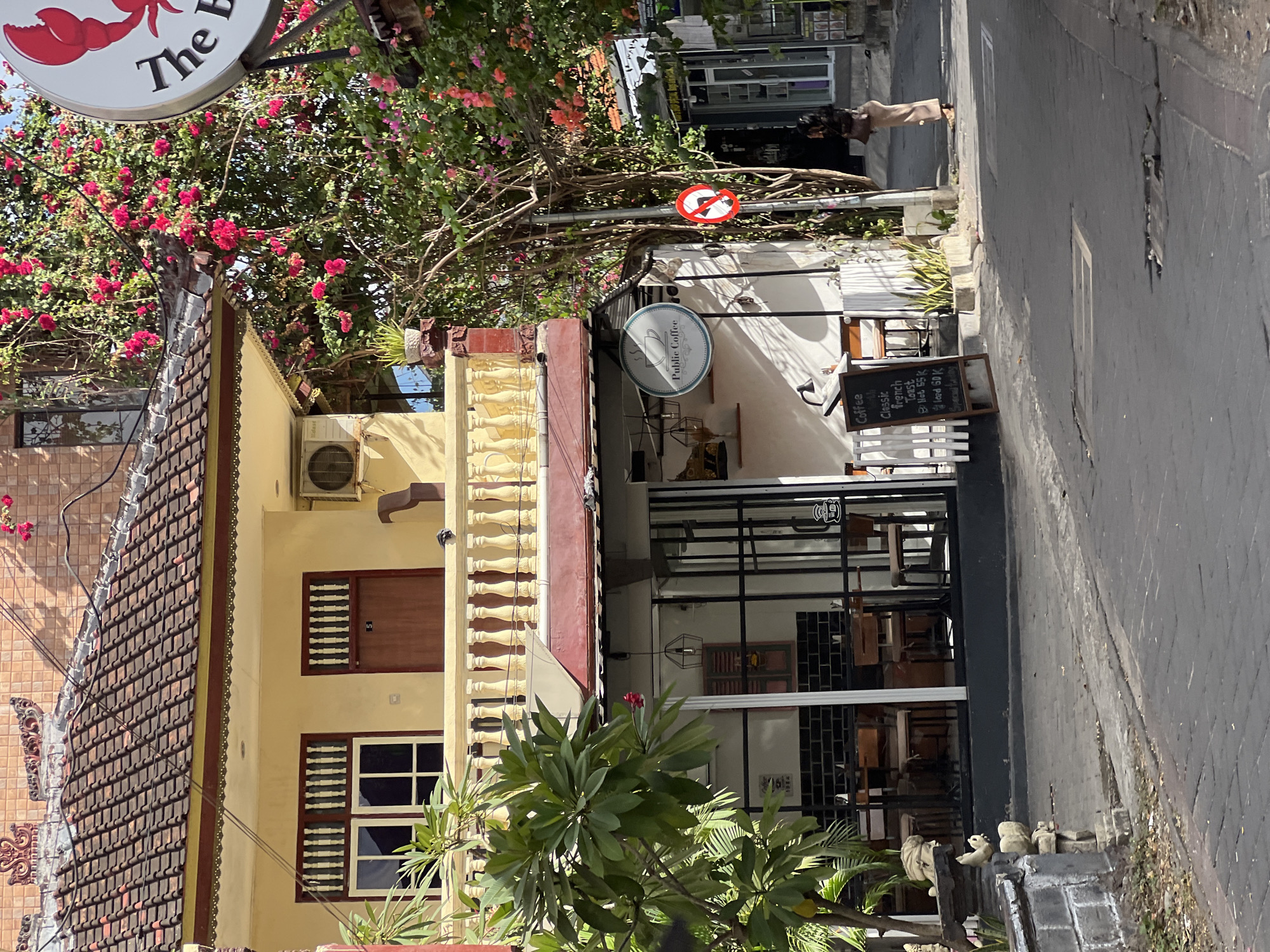 A narrow, paved street in Kuta, Indonesia, is lined with buildings and vegetation. In the foreground, to the left, a low stone wall with decorative carvings borders the street. The street is made of gray cobblestones. On the right side of the street, a woman in tan pants and a dark top stands with her back to the camera, holding a brown bag.
Further down the street, a cafe named "Public Coffee" is visible. The cafe has large glass windows revealing tables and chairs inside. A blackboard sign outside the cafe lists menu items and prices. Above the cafe, a two-story building with a yellow facade and a red-tiled roof stands. A balcony with a railing overlooks the street. Vibrant pink bougainvillea flowers bloom on trees and vines, adding color to the scene. A circular sign with a red crab and the text "The B" is visible on the right side of the image. A circular red and white sign with a diagonal line through it indicates no entry. The overall atmosphere suggests a bright, sunny day in a commercial area.