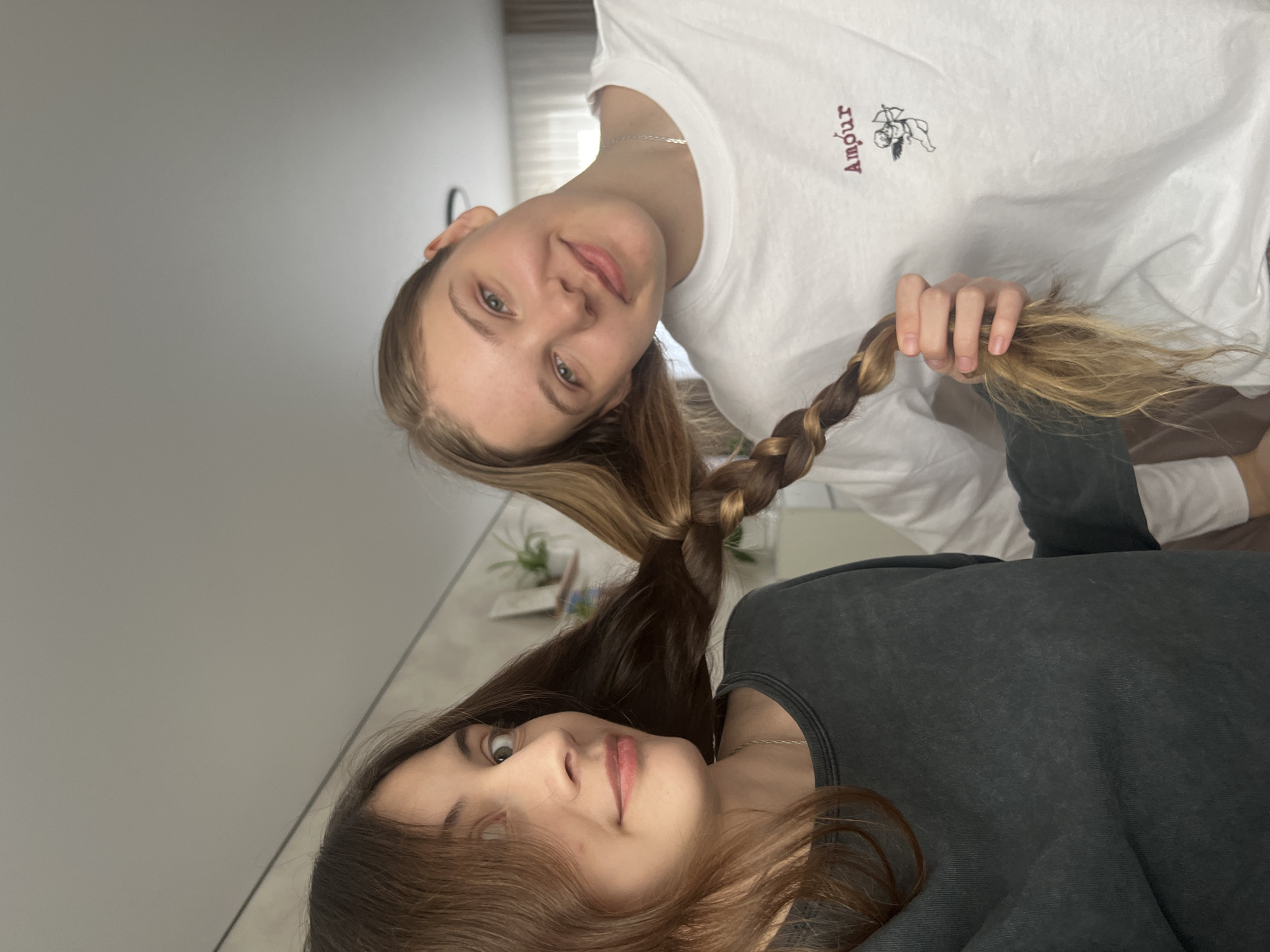 Two young women are close together and appear to be posing for a photo. One woman has fair hair, light skin, and is wearing a white, long-sleeved shirt with the word "Amour" and a small graphic on it. Her hair is partially braided. The other woman has dark hair, light skin, and is wearing a dark grey sweatshirt. Their hair is connected in a braid. The background shows part of a room with light-colored walls. The location in the city of Votkinsk, Russia, cannot be determined from the image.