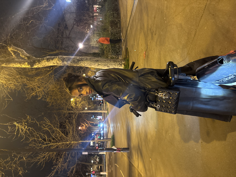 A woman in a black leather jacket is perched on a black scooter, holding a black Dior handbag. The backdrop features an urban night scene with bare trees, streetlights, and buildings. The scooter's body is visible, including a storage compartment with the words "Uber" partially visible. The focus is on the woman's face and the details of her attire and accessories. The lighting is dim, casting shadows across the scene.