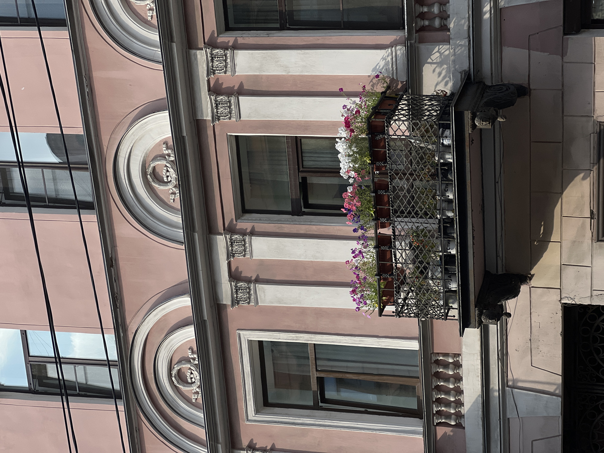The image shows a close-up view of the facade of a building in Saint Petersburg, Russia. The building is painted a pale pink color and features ornate architectural details, including arched windows with decorative moldings and fluted pilasters. A wrought-iron balcony extends from the second story, adorned with planters filled with blooming flowers in various shades of pink, white, and purple. The balcony itself is supported by decorative brackets. The sky is visible in the reflection of the windows, suggesting it is a clear day. The overall impression is of a well-maintained, historically significant urban structure.