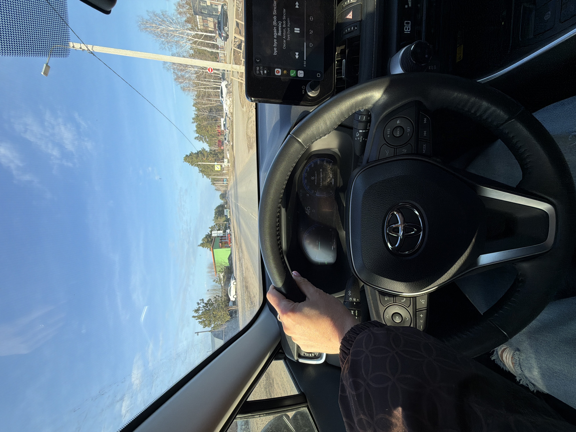 The image shows the interior of a car, specifically the driver's perspective. A hand rests on the steering wheel, emblazoned with the Toyota logo. The car's infotainment system displays the song "Bye Bye Again (Bob Sinclar Remix)" by Oscar Anton and Bob Sinclar. The speedometer reads 46, and the gear is set to 'D'. The exterior view through the windshield shows a road lined with trees and buildings. Based on the visible text and context, the location appears to be Поляны, Russia.