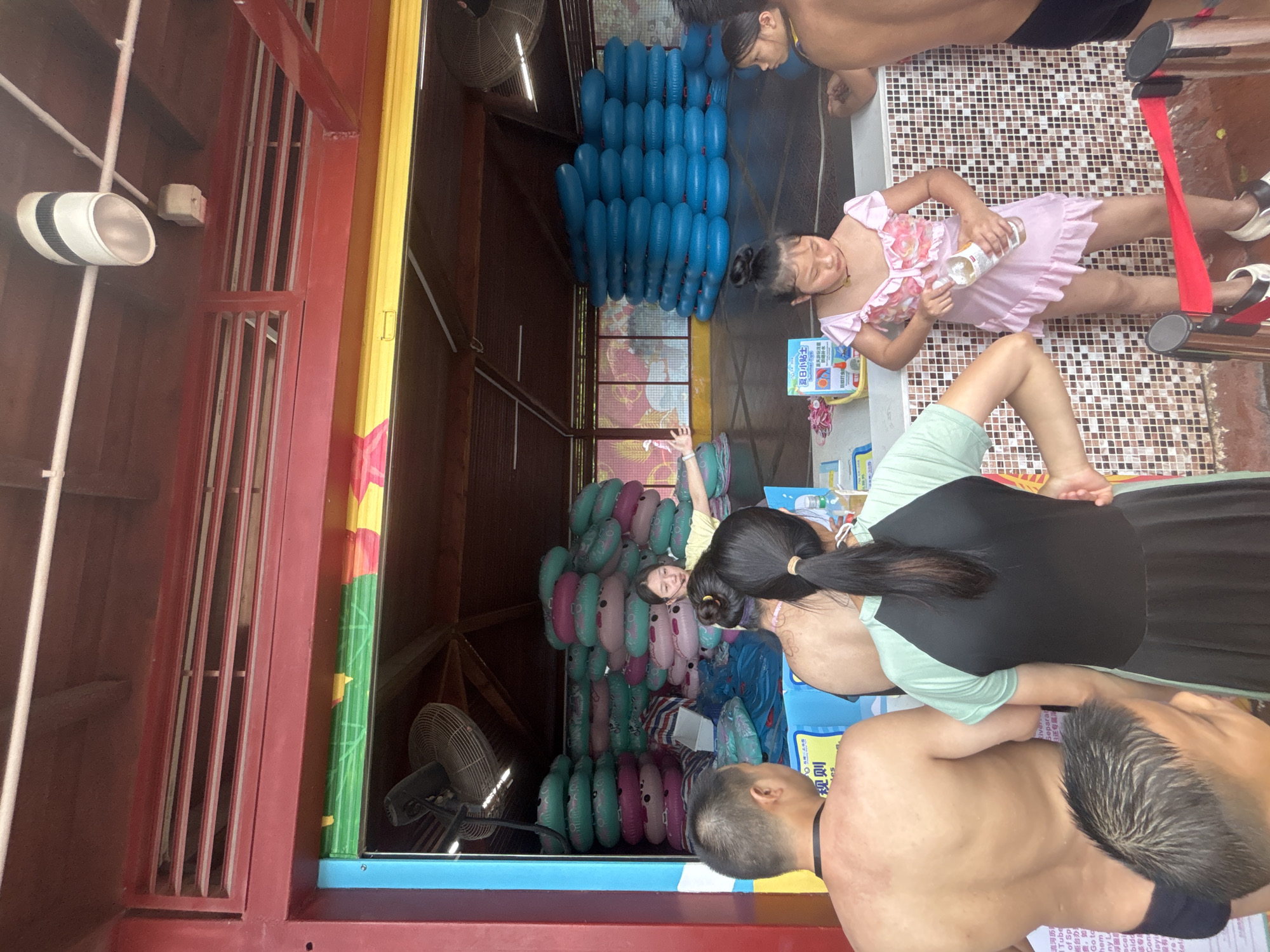 In Panyu District, China, a scene unfolds at what appears to be a water park or amusement facility. The primary focus is a service counter, constructed from red metal and topped with a yellow fascia, against a backdrop of wooden beams. Stacks of colorful inflatable tubes, predominantly teal and pink, are piled high behind the counter, suggesting their rental or sale. A young woman in a yellow shirt is actively engaged with customers, gesturing with her hands. In the foreground, three individuals are visible. To the left, a man is seen from the back, shirtless and wearing dark swim trunks. In the center, a woman in a black and teal dress stands with her back to the camera. To the right, a young girl in a pink frilly swimsuit holds a clear bottle, appearing to be drinking from it. Behind the girl, another person is partially visible, also appearing to be a customer. The environment suggests a warm climate, with an oscillating fan visible on the left, and the tiled wall behind the counter indicating a wet environment. A red rope barrier with metal stanchions is present in the lower right, likely demarcating a queue.