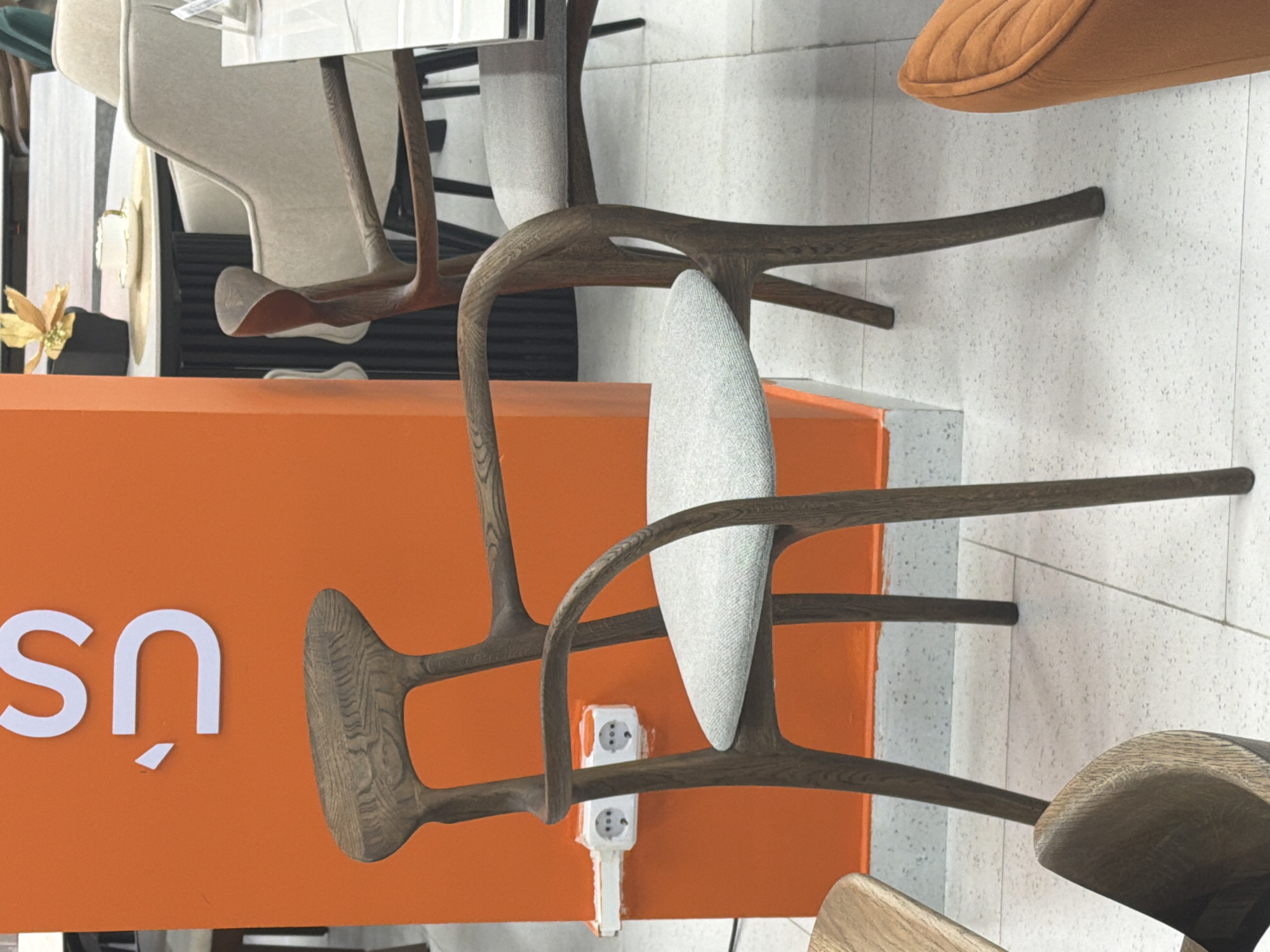 A wooden chair with a light grey upholstered seat and back is the central focus of the image. The chair has curved armrests and a distinctively shaped back. It is positioned in front of an orange wall with white electrical outlets. To the right of the chair, another similar chair is partially visible, and further back, a table with a white cup and saucer and a decorative plant can be seen. The floor is tiled with a light-colored speckled pattern. The image appears to be taken inside a furniture store or showroom. The presence of the electrical outlets and the arrangement of furniture suggest an indoor retail or exhibition space, potentially in Astana, Kazakhstan, as indicated by the provided context.