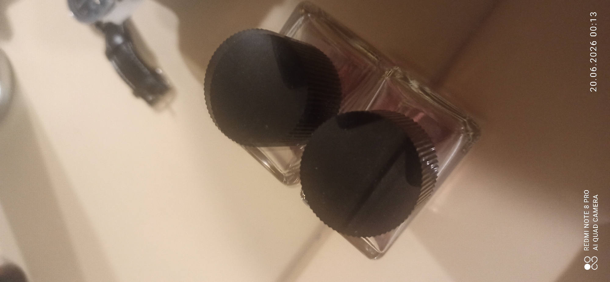 Two black screw-on caps are placed on top of a clear glass bottle with a pinkish liquid inside. The bottle is sitting on a light-colored surface that appears to be a countertop. The caps are ribbed and have a flat top with a slight indentation. The lighting in the scene is dim, creating shadows on the surface and the objects. The background is blurred and out of focus. There is a timestamp in the lower right corner indicating "20.06.2026 00:13". There is also text in the lower left corner indicating "REDMI NOTE 8 PRO AI QUAD CAMERA". There is no indication of location within a city.