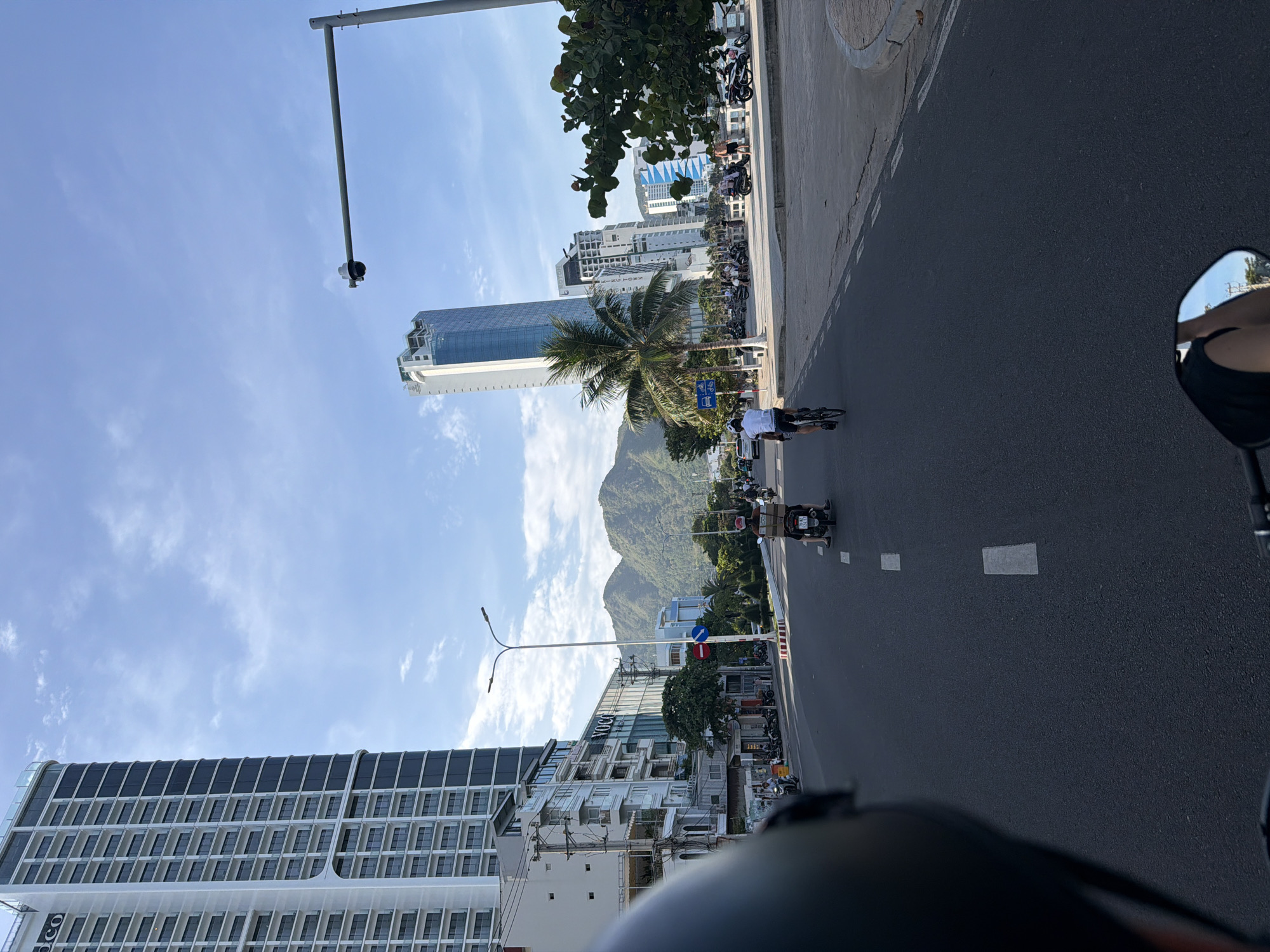 The image shows a view from a motorcycle or scooter, looking down a street in Bac Nha Trang Ward, Vietnam. A person's black helmet is visible in the foreground on the left, and a mirror reflecting the rider's arm is in the bottom right corner.
The street is paved and marked with white dashed and solid lines. Several people are riding motorcycles or scooters, and one person is riding a bicycle. One scooter rider in the middle of the frame is carrying a large box on the back. Traffic appears to be moving in the same direction as the viewpoint.
On the left side of the street, there are several multi-story buildings, including a tall, modern high-rise building with many windows. Further down the street, there are more buildings of varying heights. On the right side of the street, there is a paved walkway and a line of trees, including a palm tree. In the background, there are mountains and a clear blue sky with some scattered clouds. A traffic light and a street lamp with a surveillance camera are also visible. The scene appears to be set during daylight hours.