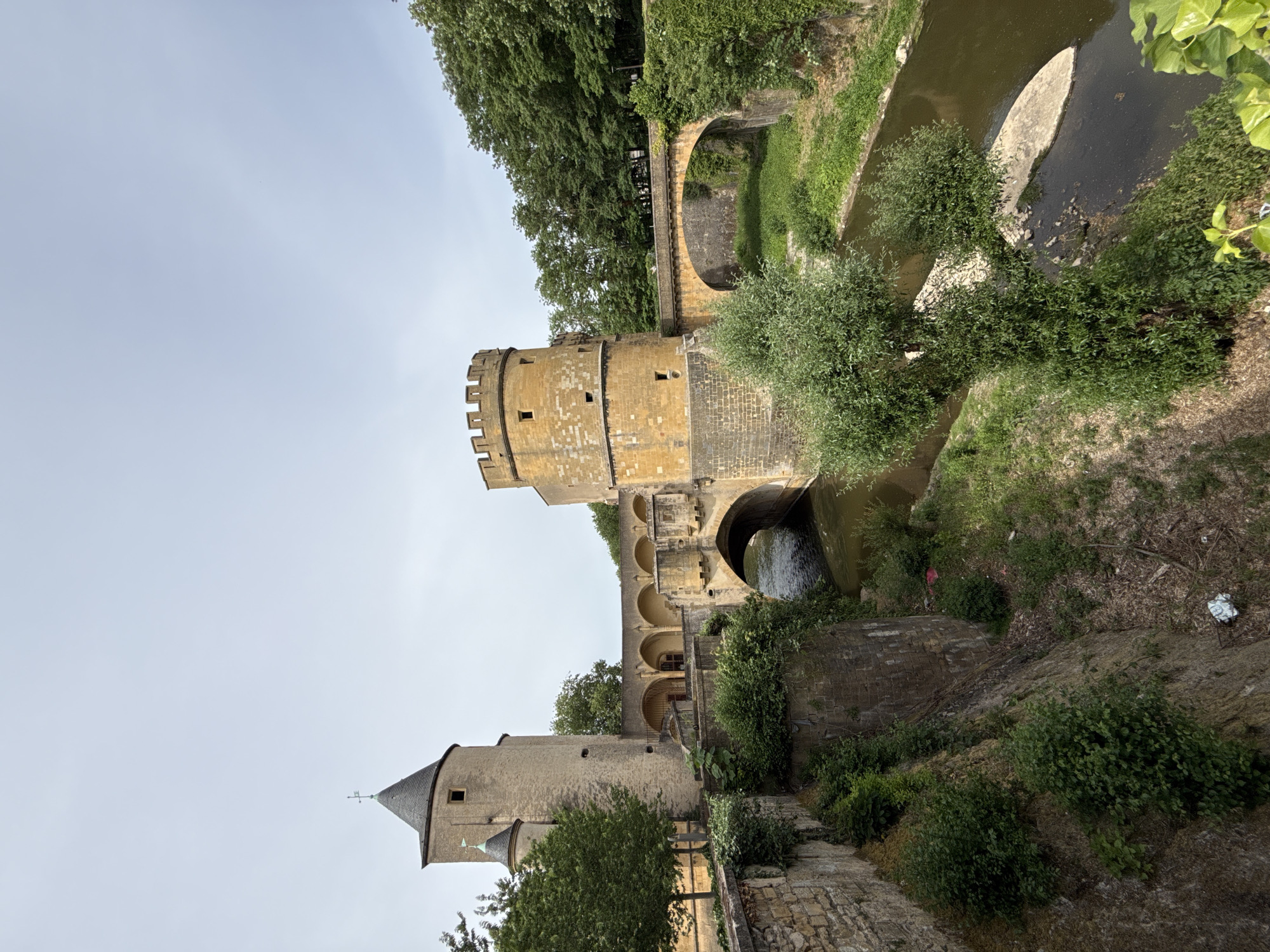 The image shows the Porte des Allemands in Metz, France. This medieval gate is constructed of yellow-brown stone and features two towers, one with a conical roof. The gate spans the Seille river, which flows through a series of stone arches. Vegetation grows on the banks of the river, including trees and bushes. The sky is overcast.