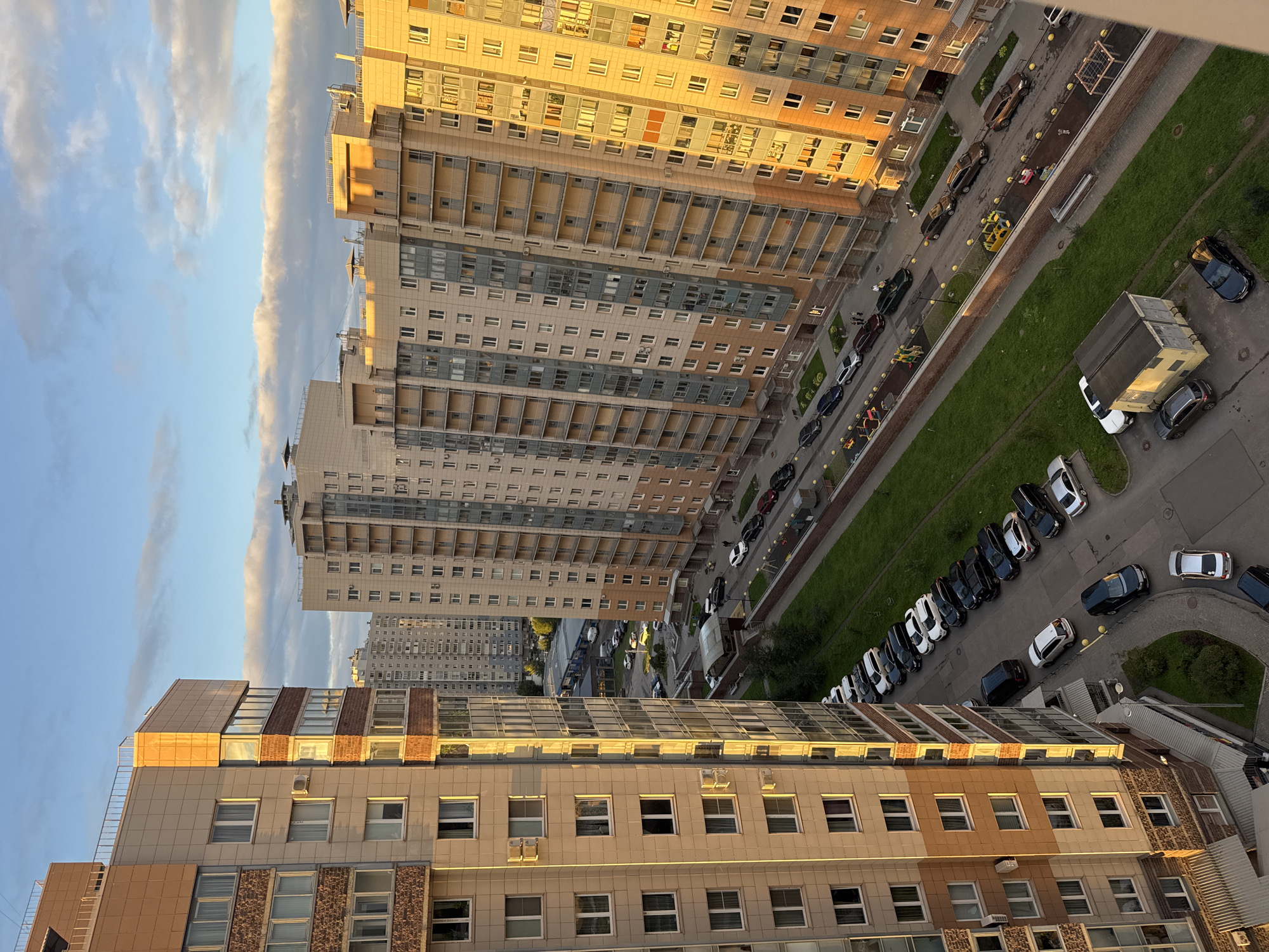 The image depicts a view of a residential area in an urban environment, likely in Russia, as indicated by the architectural style and vehicle models. The scene is dominated by tall, modern apartment buildings with multiple floors, featuring balconies and numerous windows. The buildings are constructed with light-colored facade materials, interspersed with brick or stone elements.
In the foreground, a tall apartment building on the left is illuminated by the golden light of the setting sun, casting long shadows. The building has a complex facade with varying window designs and some protruding elements.
The middle ground reveals a street lined with parked cars and a strip of green grass. Another row of apartment buildings, taller and more uniform in design, stretches into the distance. These buildings are also bathed in the warm, evening light. The street appears to be a residential road, with some playground equipment and benches visible along the side.
The background shows a clear sky with scattered clouds, suggesting it is late afternoon or early evening. The overall impression is a typical urban landscape of a residential district.
Several cars are parked along the street and in designated parking areas. The cars are of various makes and models, predominantly sedans and SUVs, suggesting everyday urban transportation. There are no people prominently featured in the scene, though small figures could be present but are not discernible at this resolution. The environment appears to be well-maintained, with green spaces and paved roads.