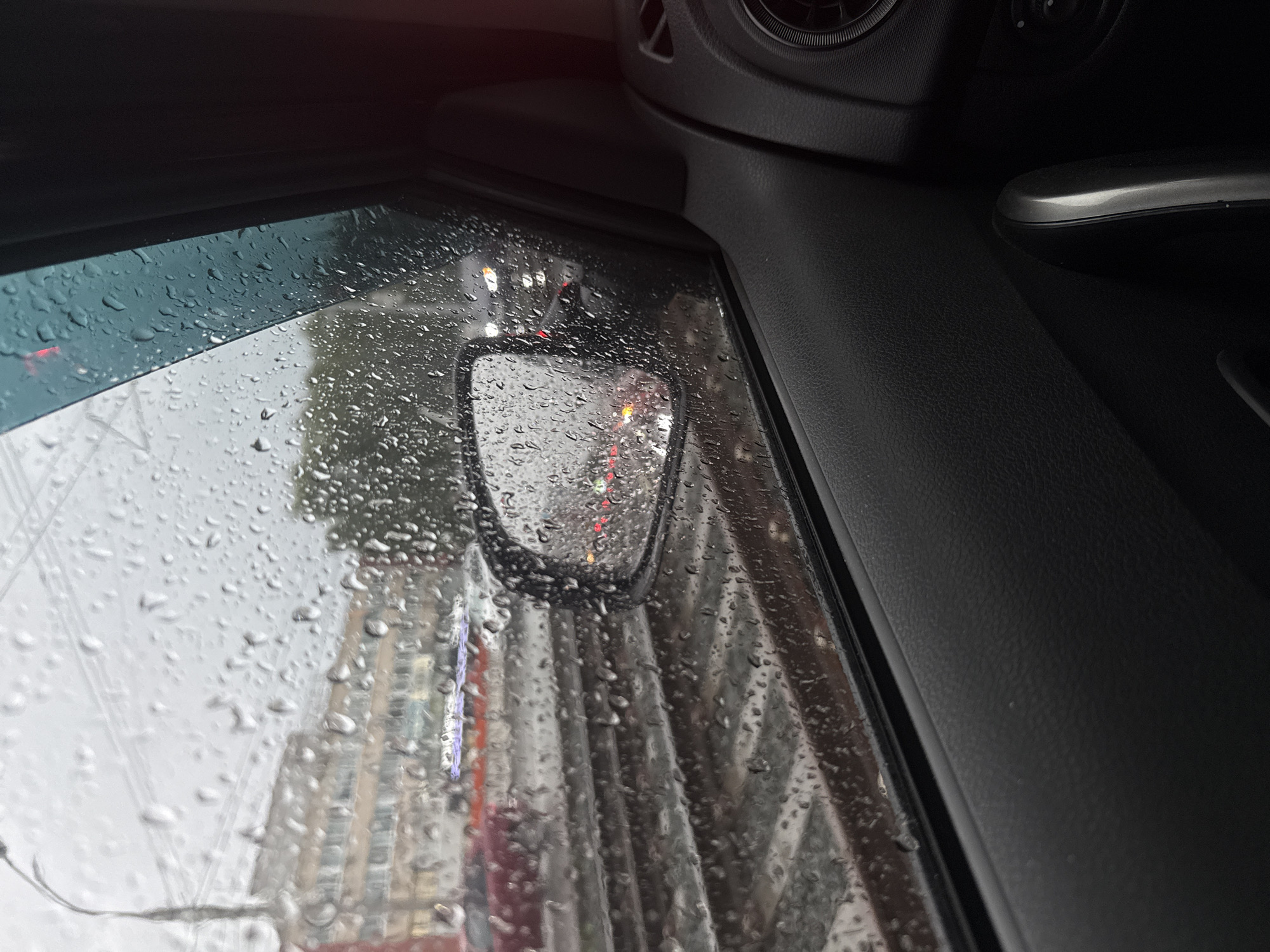 The image shows a view from inside a car on a rainy day in Izhevsk, Russia. Water droplets cover the car window, obscuring the view of the city outside. A small rectangular side mirror is visible on the car's door, providing a blurred reflection of the street scene. The car's interior, including the dashboard and door panel, is visible in the foreground.