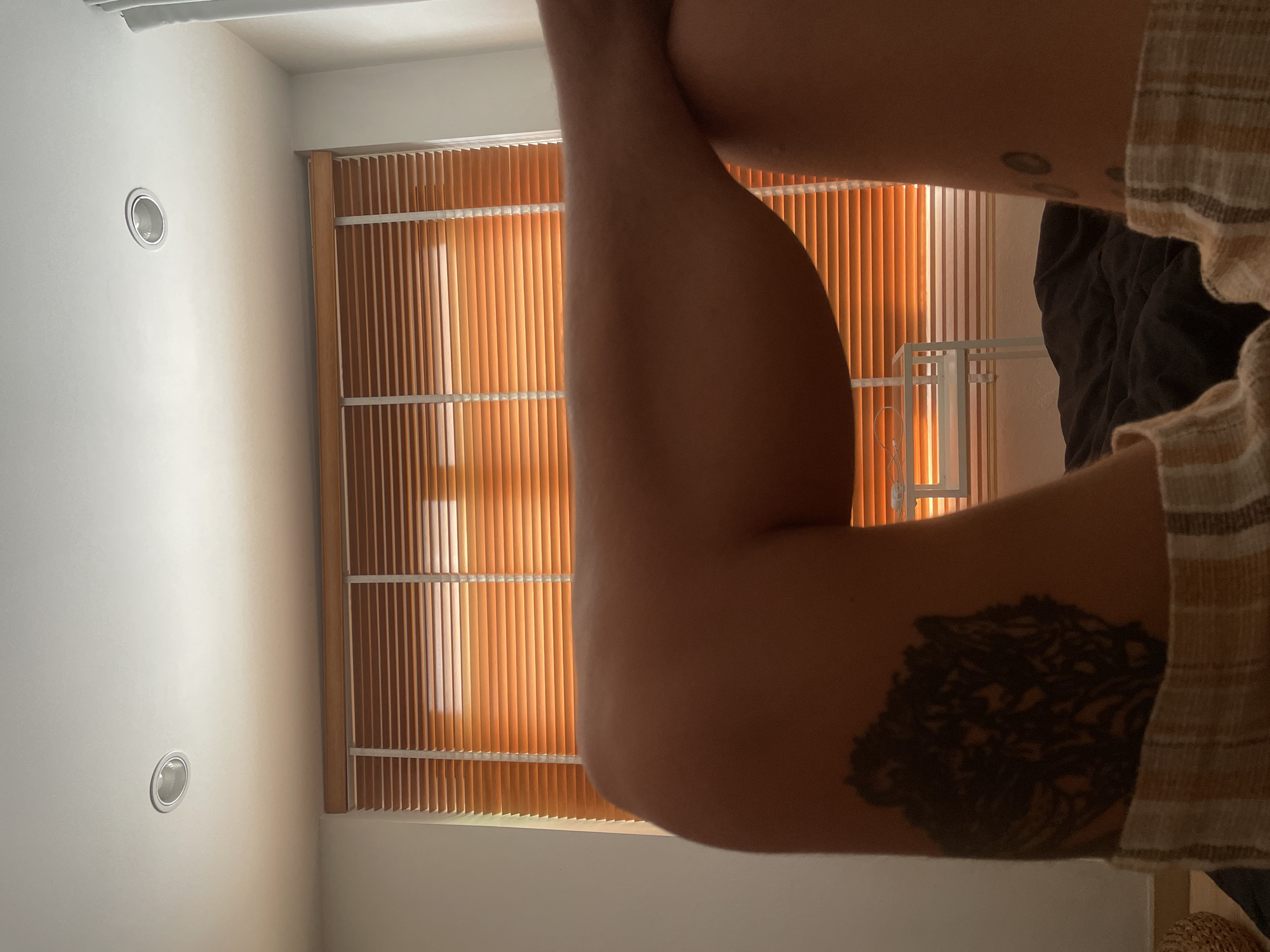 A close-up, indoor shot features a person's bent arm with a tattoo on the forearm. The arm is positioned against a window covered with closed, orange-toned vertical blinds. A white wall is visible to the left of the window. A portion of the room with a dark blanket and a white metal frame can be seen on the right. The location is possibly within a building in Chang Phueak, Thailand.
