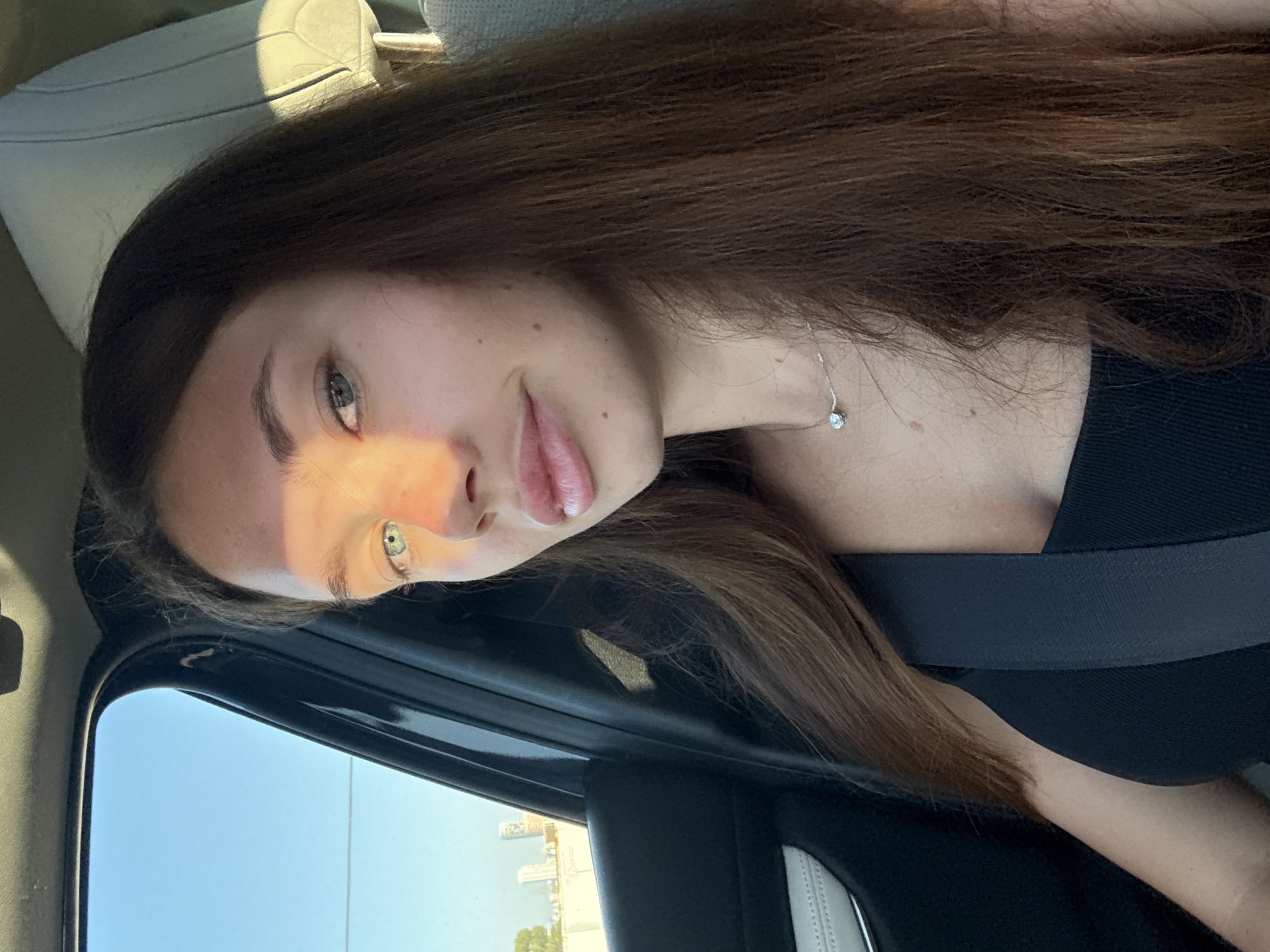 A young woman with long brown hair is in a car, wearing a black top and seatbelt. She is looking at the camera, and the sun is shining on her face. Outside the car window, a cityscape, possibly of Tel Aviv, Israel, is visible on the coast.