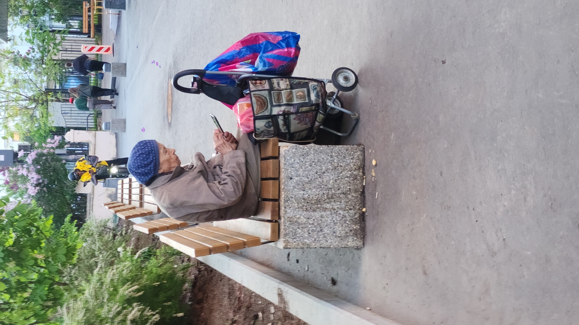 An older woman sits on a bench outdoors in Moscow, Russia. She is wearing a blue headscarf and a tan jacket, and she is looking at her cell phone. Beside her is a shopping cart with a patterned bag and a blue and pink striped bag. In the background, a person in yellow rides a scooter, and other pedestrians walk by. The area has trees and flowering bushes. The scene appears to be captured during the daytime with natural lighting. The image is rotated sideways.