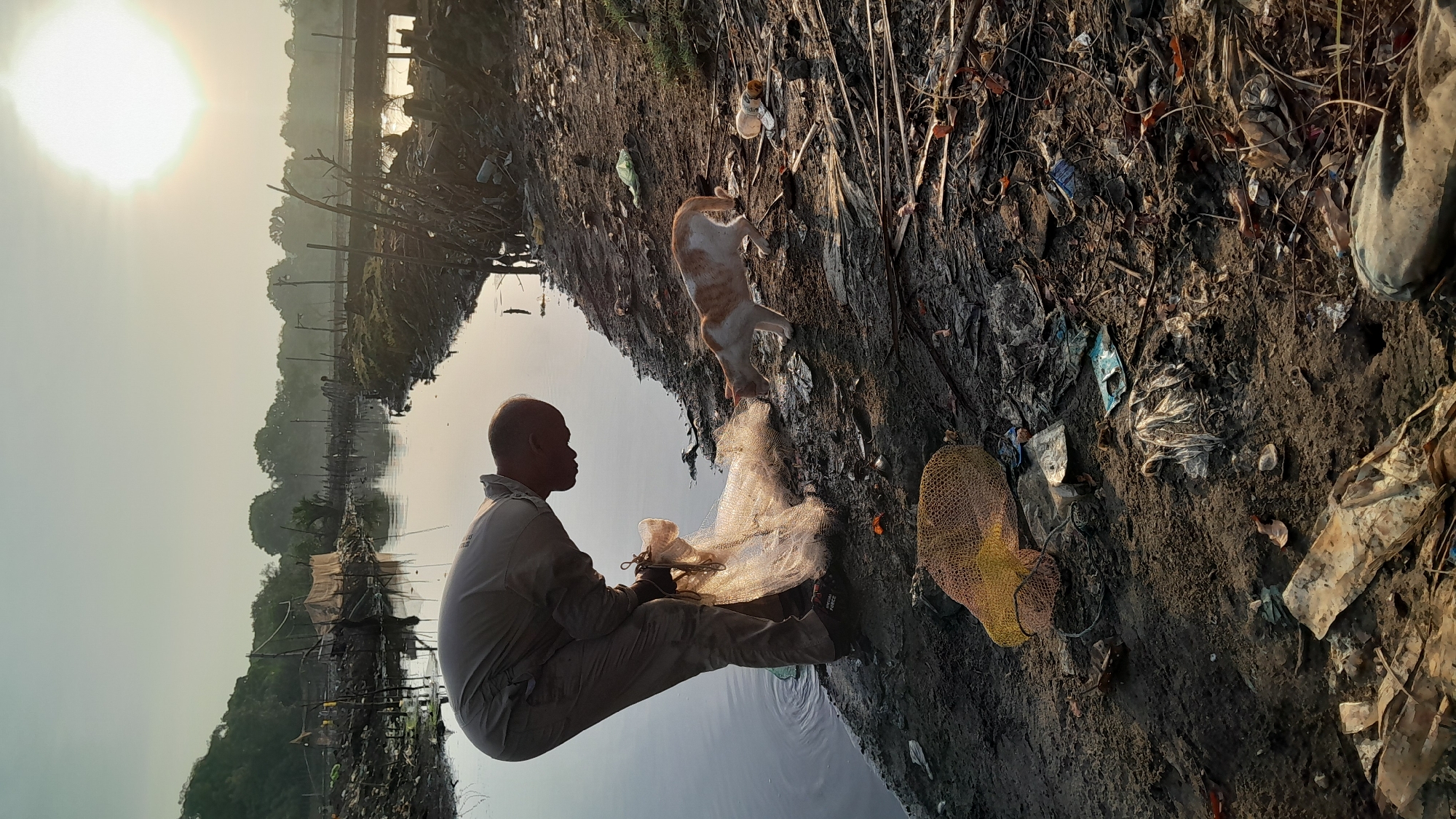 An adult male is depicted bent over, engaging with a white, fine-mesh fishing net partially submerged in a polluted waterway in Lhokseumawe, Indonesia. He wears light-colored overalls and dark shoes, with "SPECIAL FORCE" visible on his right shoe. An orange and white cat stands on the muddy bank to his right, sniffing the ground near the net. The immediate bank area is heavily littered with various refuse, including plastic bottles, bags, fabric scraps, and debris. A yellow mesh bag is also present on the ground. The background shows the waterway extending under a hazy morning sky, with the sun appearing as a bright, diffuse orb. The far bank features informal structures, dense vegetation, and utility poles. Ripples are visible on the water's surface near the man's activity.