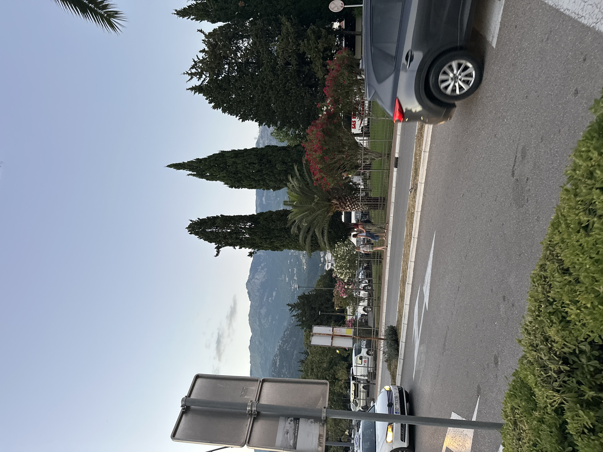 The image depicts a street scene in Budva, Montenegro, with a view of the mountains in the background. In the foreground, a gray SUV is driving on the right side of the road. A white car, possibly a taxi with a license plate BD TX375, is parked on the left. A traffic sign is visible on the left side of the image. Several people are walking on the sidewalk. Tall cypress trees and flowering bushes line the street. The sky is clear with some clouds.