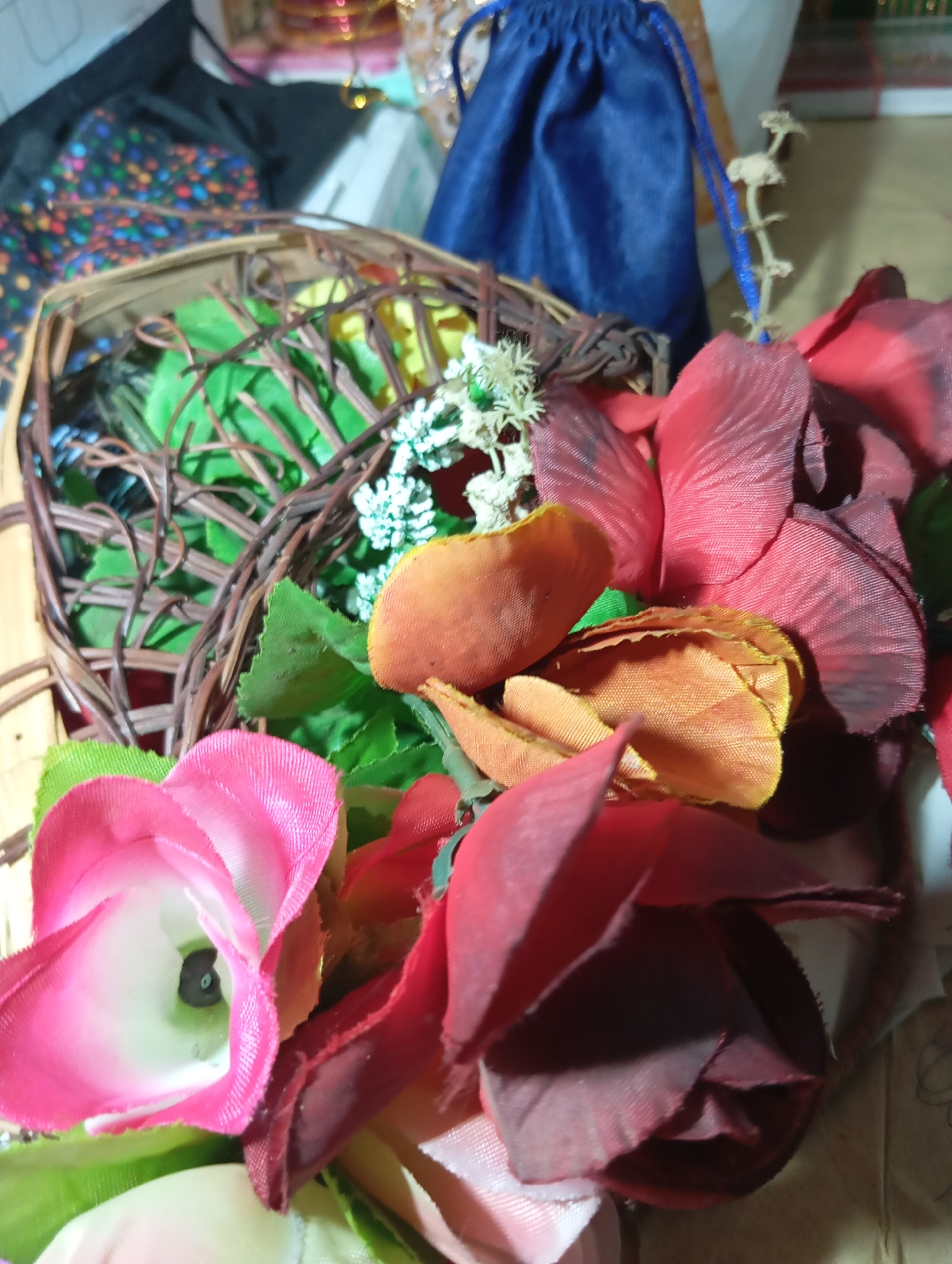 A close-up, overhead shot shows an assortment of artificial flowers arranged in and around a small wicker basket. The flowers are in various shades of pink, red, and orange, with some green foliage visible. In the background, a dark blue drawstring bag and a patterned fabric are partially visible, suggesting an indoor setting. The lighting is dim, with a slight yellow cast.