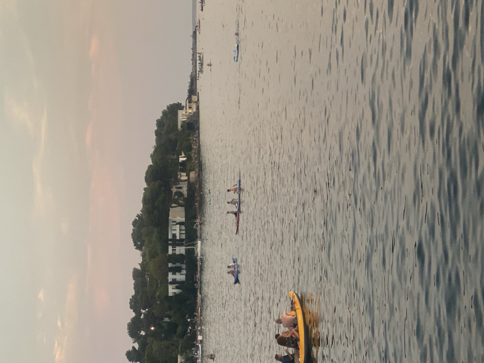 The image depicts a serene sunset scene over the water in Antibes, France, with a shoreline populated by trees and buildings. In the foreground, a yellow kayak with four individuals is visible. Further out, another kayak with two people is observed. In the mid-ground, several other kayaks and small boats are scattered across the water, with some individuals engaged in paddling. The background reveals a stretch of coastline characterized by lush greenery and white buildings, suggesting a residential or resort area. A sailboat is also present near the shore. The sky is adorned with soft, pastel-colored clouds, indicating the twilight hour. The water is calm with gentle ripples.