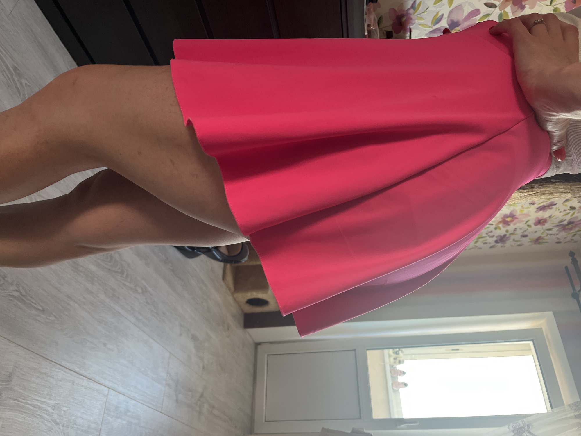 The image captures a close-up view of a person from the waist down, wearing a vibrant pink skater skirt. The skirt is flared and appears to be made of a light, possibly synthetic material, with a clean hemline. The person's legs are visible below the skirt, and they appear to have tanned skin with some minor marks or blemishes. The background suggests an indoor domestic setting, with a dresser or cabinet on the left side of the frame and a floral wallpaper pattern behind it. To the right, a white-framed window is visible, with light streaming in from outside, and a glimpse of a balcony or outdoor area beyond. A cat tree or scratching post is partially visible in the mid-ground. The flooring appears to be laminate or wood-look tiles. The image is taken from a low angle, emphasizing the skirt and legs.