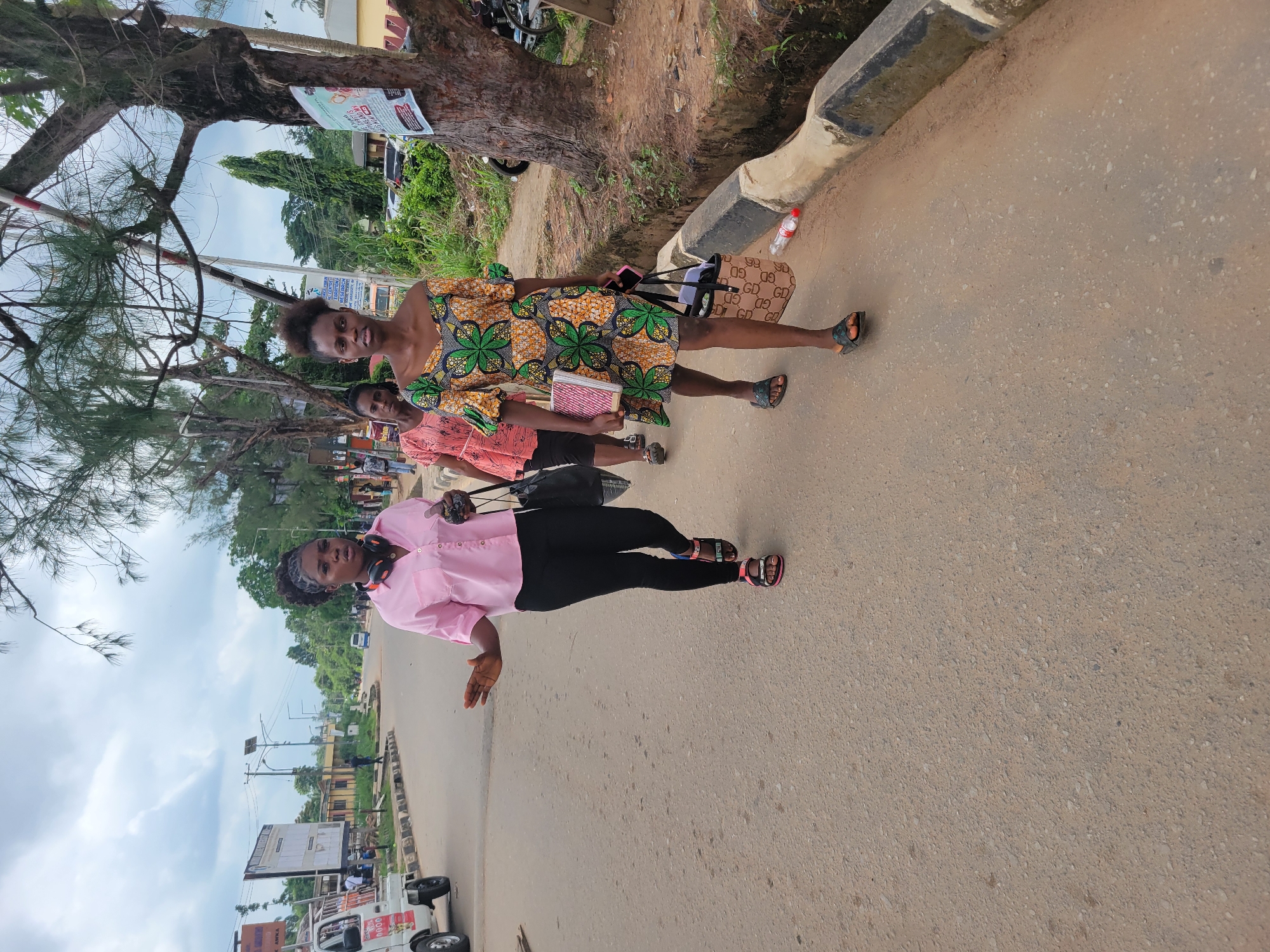 Three young women are walking on a paved road in Egbagu, Nigeria. The woman in the foreground is wearing a short, brightly colored green and orange patterned dress and black sandals. She is carrying a patterned tote bag and a pink mobile phone. The woman in the middle is wearing a pink short-sleeved shirt, black pants, and black sandals with pink accents. She has headphones around her neck and is carrying a black tote bag. The woman in the background is wearing a light orange, short-sleeved dress. The road is a light brown asphalt with a concrete curb and a shallow drainage ditch on the right side of the image. Trees line the right side of the road, with foliage and some taller trees in the background. The sky is overcast. In the background, there are buildings, streetlights, utility poles, and vehicles, including a white truck on the left.