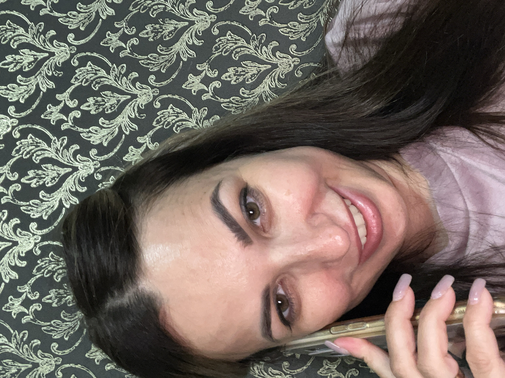 A woman with dark brown hair and brown eyes is holding a smartphone to her left ear and smiling. She is wearing a light purple t-shirt. The background is a dark wallpaper with a repeating floral pattern in a lighter color. The lighting is artificial and appears to be coming from the front.