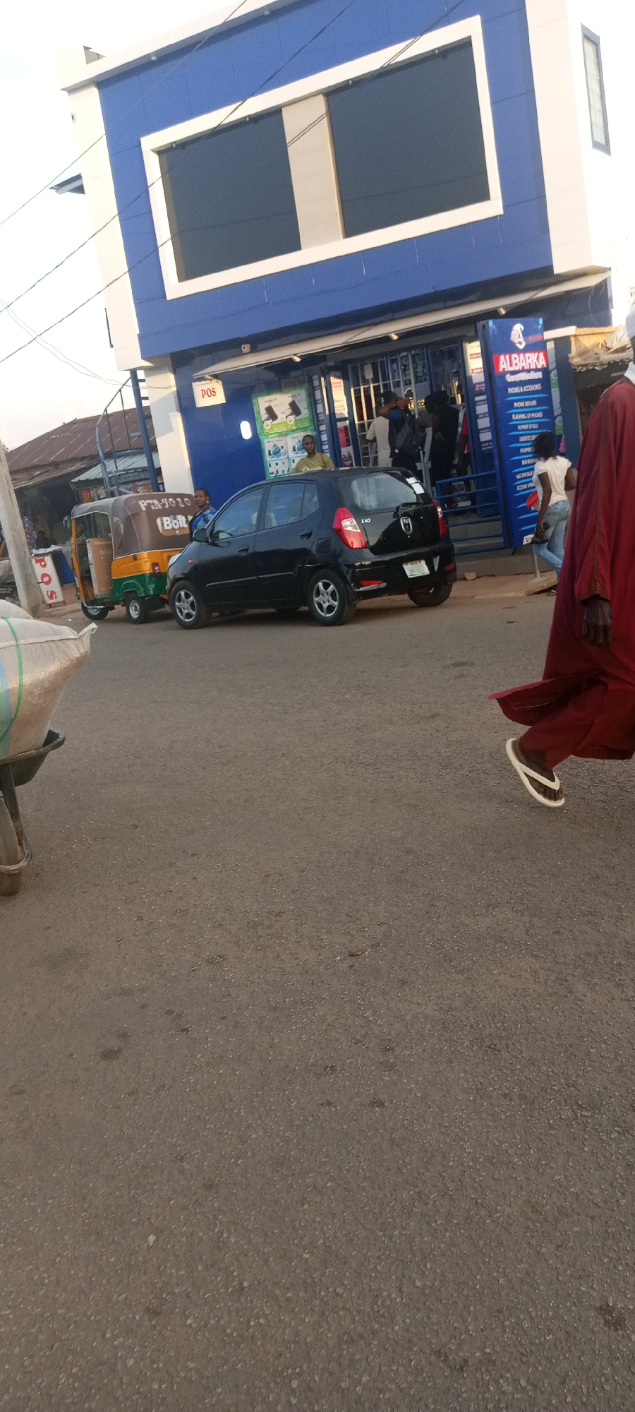 The image depicts a street scene in what appears to be Jos, Nigeria. In the foreground, the lower portion of a man wearing a red robe is visible, suggesting he is in motion. The street surface is asphalt. In the middle ground, a black Hyundai i10 car is parked near the curb of a blue and white two-story building. A yellow and green tricycle, often used for transport in Nigeria, is parked further back. People are gathered around the building, which has a sign indicating it is a business named "ALBARKA." The sign lists "Phones & Accessories," "Phone Repairs," and "Flashing of Phones," suggesting it's a phone service shop. A "POS" sign is visible above the entrance. A wheelbarrow with bags of materials is on the left foreground. Overhead power lines are visible. The general environment is urban, with a mix of commercial and residential elements.