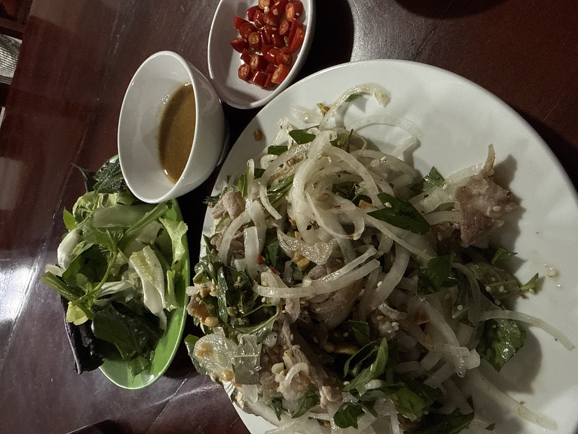 The image displays a close-up, overhead view of a Vietnamese meal served on a dark wooden table. The main subject is a white plate containing a salad made with shredded meat, thinly sliced onions, and fresh herbs, possibly Vietnamese coriander and mint, mixed with what appears to be crushed peanuts and a light dressing. To the right of this salad is a small white dish filled with chopped red chilies. Behind the main plate, a green plate holds a pile of mixed fresh greens, including leafy vegetables and possibly some cucumber slices. Further back and to the right, a small white bowl contains a dipping sauce, likely fish sauce-based, common in Vietnamese cuisine. The lighting is dim, suggesting an evening or indoor setting. The image is a still life of food, with no people or actions depicted. Based on the presence of fresh produce and the style of the dish, it is plausible that this meal was prepared and consumed in the city of Da Lat, Vietnam, known for its agricultural produce and fresh ingredients.