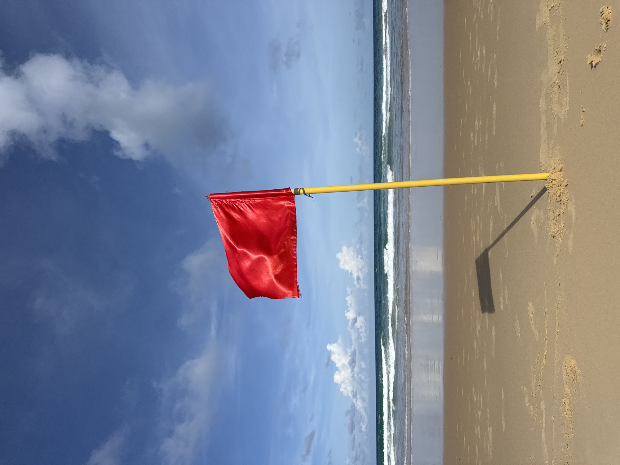 A solitary red flag waves on a yellow pole on a sandy beach. The flag is a bright red rectangle, positioned to the left of the frame. It is attached to a thin, yellow pole that is firmly planted in the sand. The beach itself is a light tan color, with subtle ripples and footprints visible in the sand. In the background, the turquoise ocean meets the shore with gentle waves. The sky above is a vibrant blue, dotted with fluffy white clouds. The scene appears calm and serene, suggesting a beautiful day at the coast. This location is the beach of Karon, Thailand.