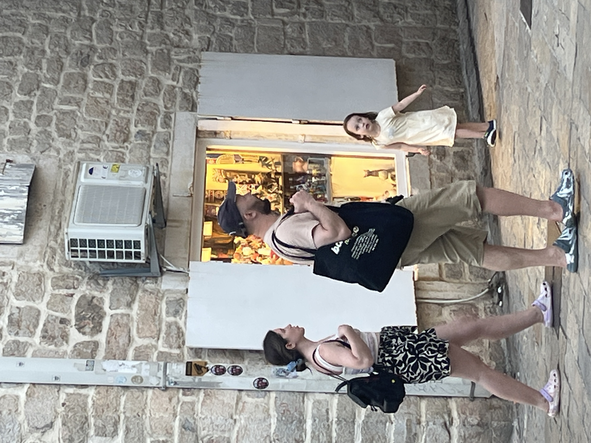 The image depicts a street scene in Budva, Montenegro. In the foreground, a woman and a man are walking past a storefront. The woman, on the left, is wearing a white tank top, patterned shorts, and light pink slip-on shoes. She has a black backpack slung over her left shoulder. The man, to her right, is wearing a light brown t-shirt, khaki shorts, a baseball cap, and patterned slip-on shoes. He is carrying a large black tote bag. Behind them, to the right, a young girl in a light yellow dress and dark sneakers is standing and looking to her left. The storefront behind the people has its shutters open, revealing merchandise displayed inside, illuminated by warm lighting. The building's facade is constructed of stone blocks, and an air conditioning unit is mounted on the wall above the storefront. The ground is paved with cobblestones.
