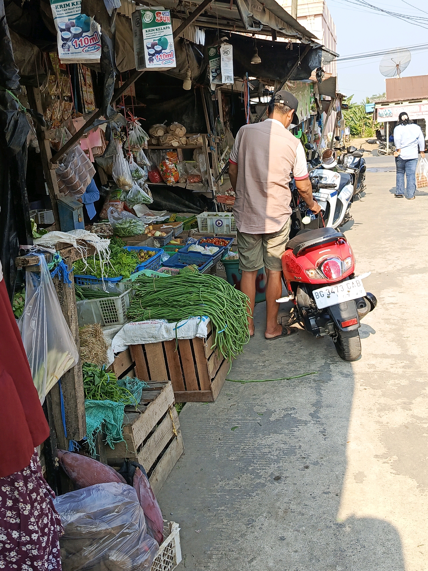 In the city of Megang Sakti I, Indonesia, a street-side market stall displays a variety of fresh produce, including bundles of long beans and leafy greens. A man wearing a light brown shirt and khaki shorts, with a black baseball cap, stands next to a red scooter, facing away from the camera. Another person, a woman in a white shirt and blue jeans, walks away from the stall, carrying a plastic bag filled with items. The stall is constructed from wood and metal, with various items hanging from the roof and shelves stocked with goods. The ground is paved with concrete, and the scene is illuminated by bright sunlight, casting shadows. A satellite dish is visible on a building in the background, suggesting a residential or commercial area.