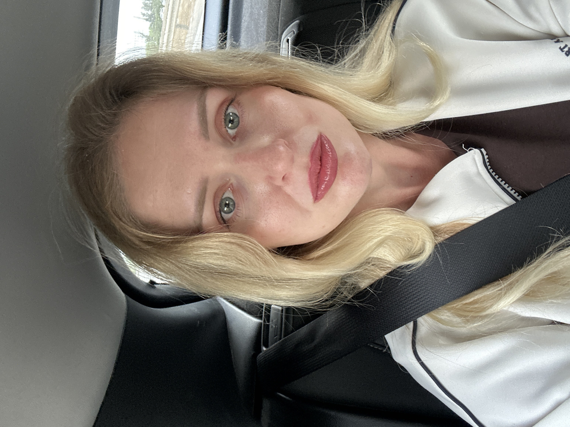 A woman with blonde hair and green eyes is in a car wearing a seatbelt and a white jacket with black trim. The interior of the car is black. The location is likely Petah Tikva, Israel, based on the request, but there are no visible details in the image confirming this.