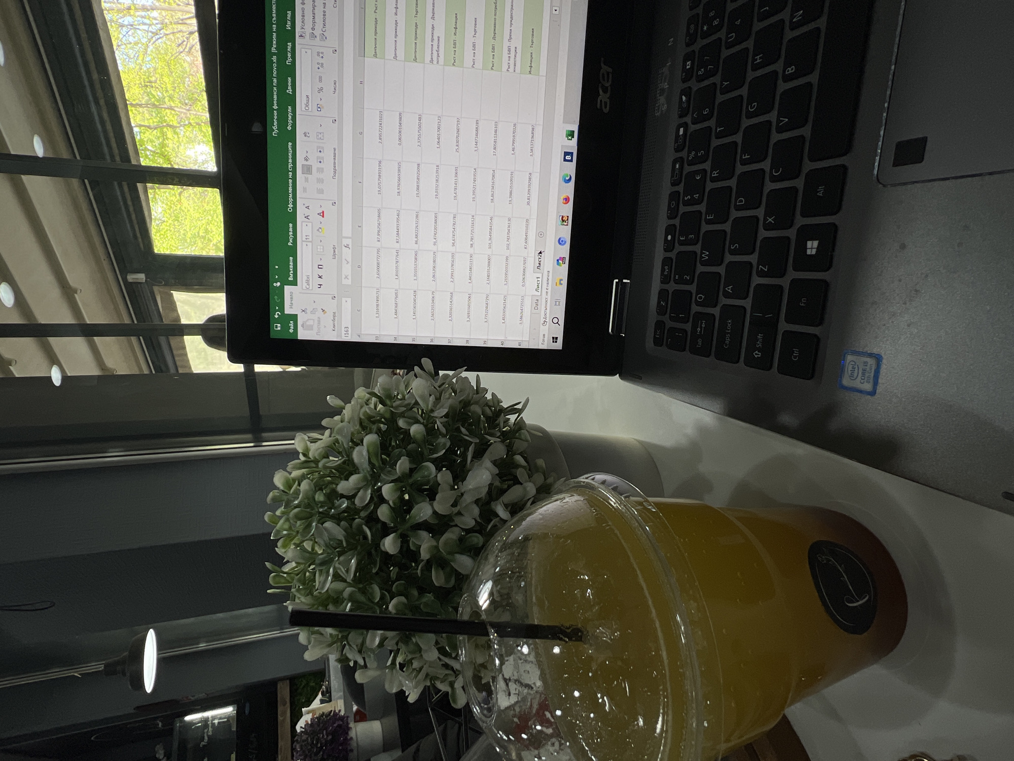 The image captures an indoor scene, likely a cafe or office in Plovdiv, Bulgaria, with a focus on a laptop, a beverage, and a small plant. The setting is bright and appears to be daytime, with ample natural light filtering through a large window, revealing lush green trees outside.
The primary subject is a silver Acer laptop, open and displaying an Excel spreadsheet. The screen shows data and menu options in Bulgarian, including "Публични финанси паi novo.xls" (Public finances on novo.xls), "Файл" (File), "Начало" (Home), "Вмъкване" (Insert), "Данни" (Data), and sheet names like "Data" and "Лист1" (Sheet1). Column headers visible include "Данъчни приходи" (Tax revenues), "Инфлация" (Inflation), and "Ръст на БВП" (GDP Growth). The laptop's keyboard has a standard QWERTY layout with some Bulgarian characters on the number keys, and an "Intel Core i5 8th Gen" sticker is visible near the touchpad.
Next to the laptop is a clear plastic cup filled with a bright yellow or orange liquid, likely an iced drink, with a black straw. The cup features a dark, circular sticker bearing a stylized white logo resembling a letter 'J' or 'T' with a swirl. Behind the drink, a small, artificial plant with numerous green and white leaves in a white pot adds a touch of decor.
The scene suggests a person is engaged in work or study, utilizing the laptop, possibly taking a break with the refreshing drink. The presence of green trees outside indicates pleasant weather. The overall ambiance is one of focused activity in a comfortable, well-lit environment.