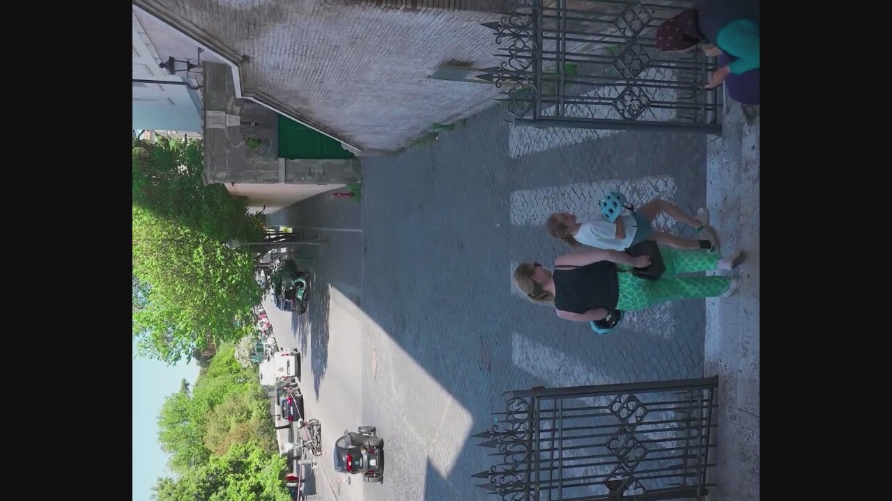 In an urban setting, possibly Castelnuovo di Porto, Italy, a woman and a girl are walking up a crosswalk. The woman, on the left, wears a black tank top and green patterned pants. She carries a dark bag in her right hand and a blue helmet in her left. The girl, to her right, wears a white t-shirt and blue shorts, carrying a blue helmet. Both are wearing sneakers. They are walking past a black metal gate. To the right of the gate, a person wearing a headscarf and a teal jacket is seated. In the background, cars and scooters are parked along a street lined with trees and buildings. A small, three-wheeled vehicle is also visible on the street. The scene appears to be on a sunny day.