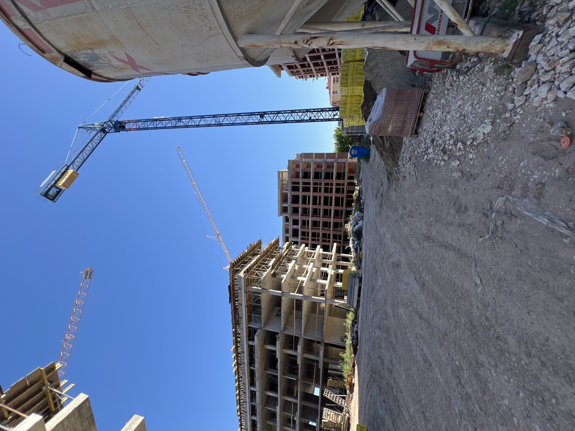 This is a photograph of a construction site in Plovdiv, Bulgaria, under a clear blue sky. In the foreground, a large, cylindrical concrete silo stands on the right, partially obscuring the view of other elements. The ground is a mix of dirt, gravel, and sand, with tire tracks visible, suggesting vehicle activity. Various construction materials are scattered around, including piles of sand, bricks wrapped in plastic on a pallet, and some wooden formwork.
Several unfinished buildings are present. The most prominent is a multi-story concrete structure in the mid-ground, appearing to be in the early stages of construction, with exposed rebar and scaffolding. To the right of this building, another structure, clad in red brick, is also under construction, with empty window frames. In the distance, additional buildings in similar states of development are visible.
Towering over the scene are two construction cranes. A large blue crane is positioned towards the center of the image, its jib extended. A second, orange crane is visible in the upper left, its jib also extended. The presence of these cranes, along with the unfinished buildings and materials, clearly indicates an active construction project. No people are visible in the frame. The overall impression is one of ongoing development within the urban landscape of Plovdiv.