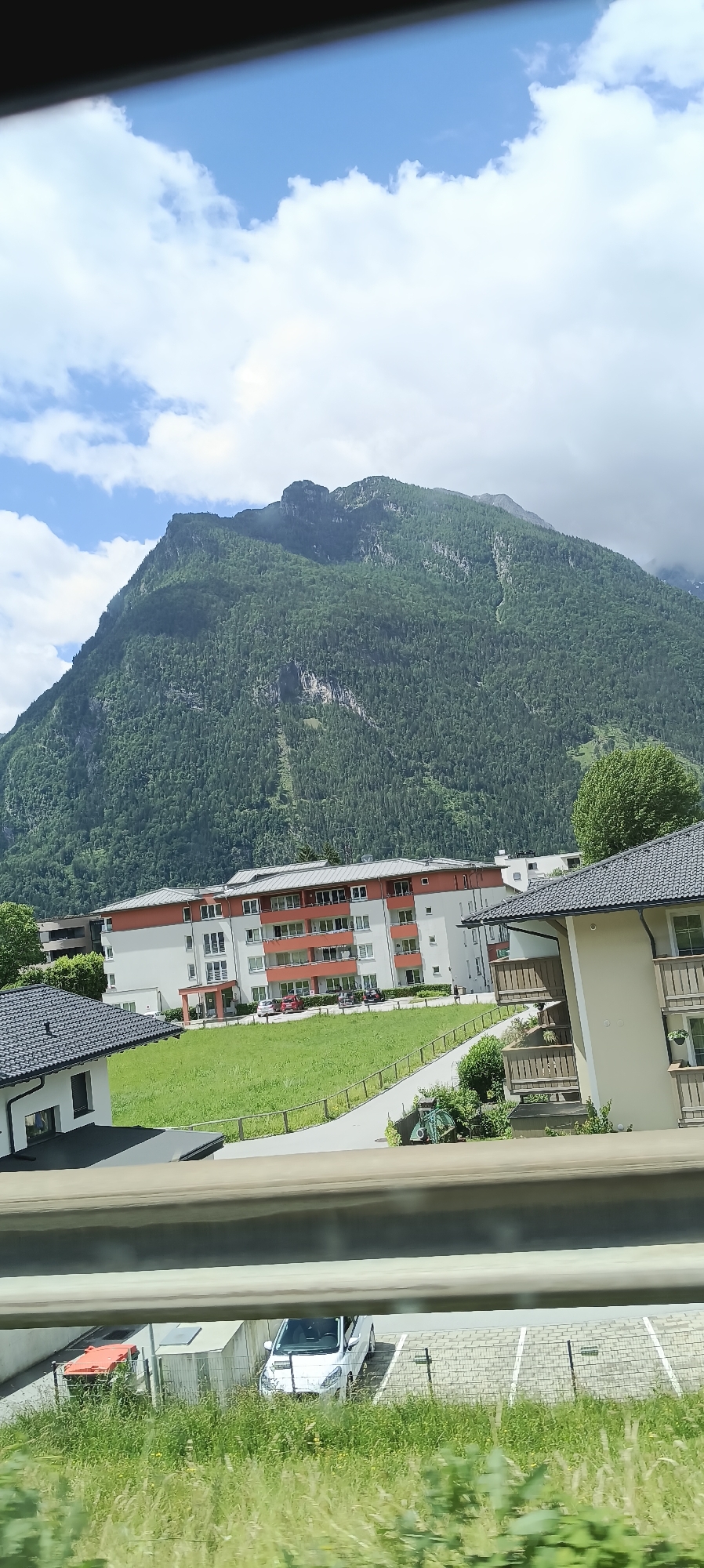 A scenic, eye-level, medium shot captures a residential area in Torren, Austria, with a majestic mountain looming in the background. The mountain is covered in dense green forest, with hints of rocky outcrops and a light dusting of clouds. In the foreground, a modern apartment building with white and orange accents stands prominently. In front of the building, a well-maintained green lawn is visible, with a paved walkway winding through it. To the right of the apartment building, another residential structure with balconies is partially visible. Below this, a parking area contains a white car and an orange dumpster. The overall scene is bathed in natural daylight, suggesting it was taken on a partly cloudy day.